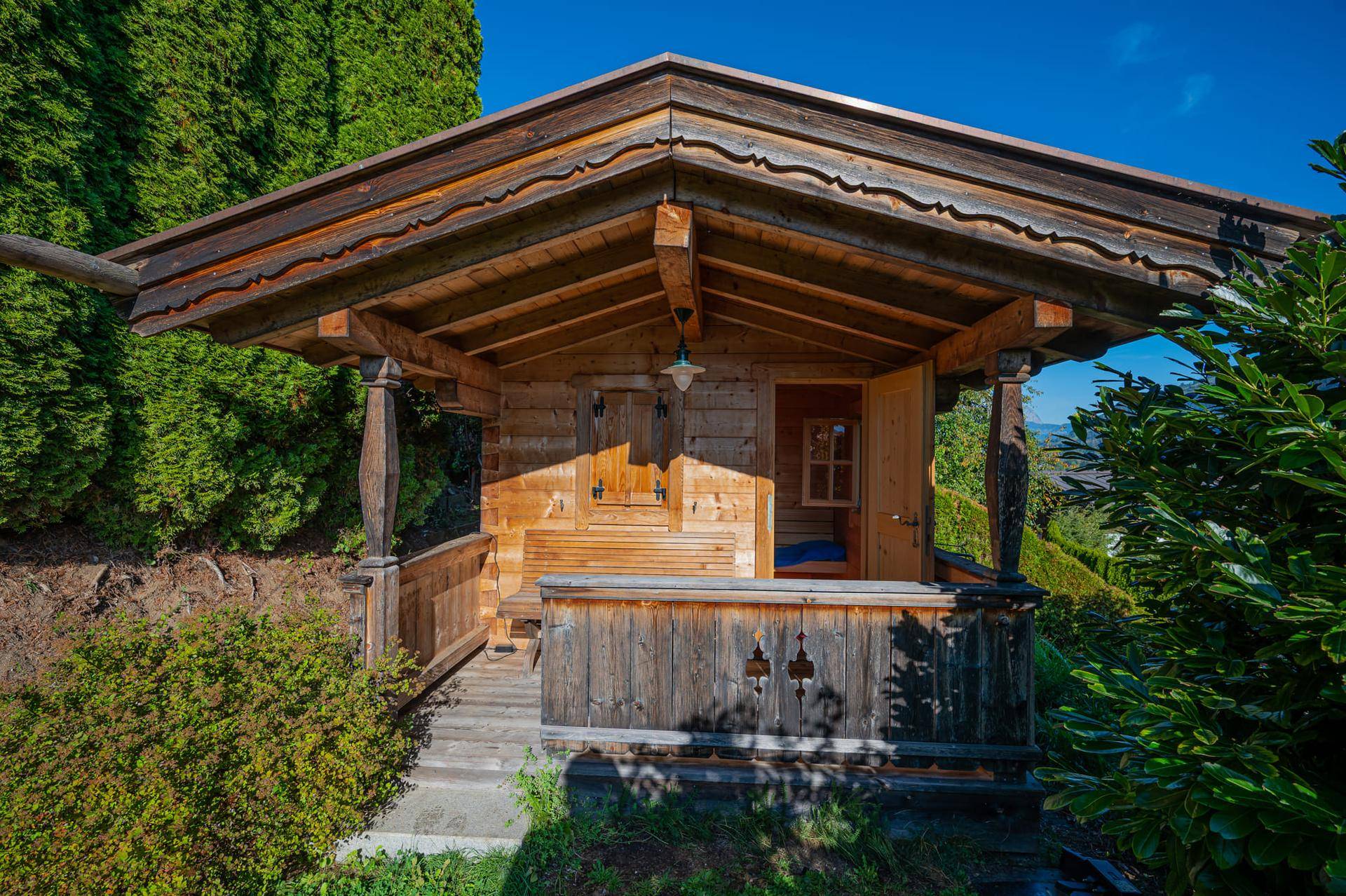 Charmantes Holzhaus im Garten mit kleiner Veranda, umgeben von üppigem Grün und Bäumen.