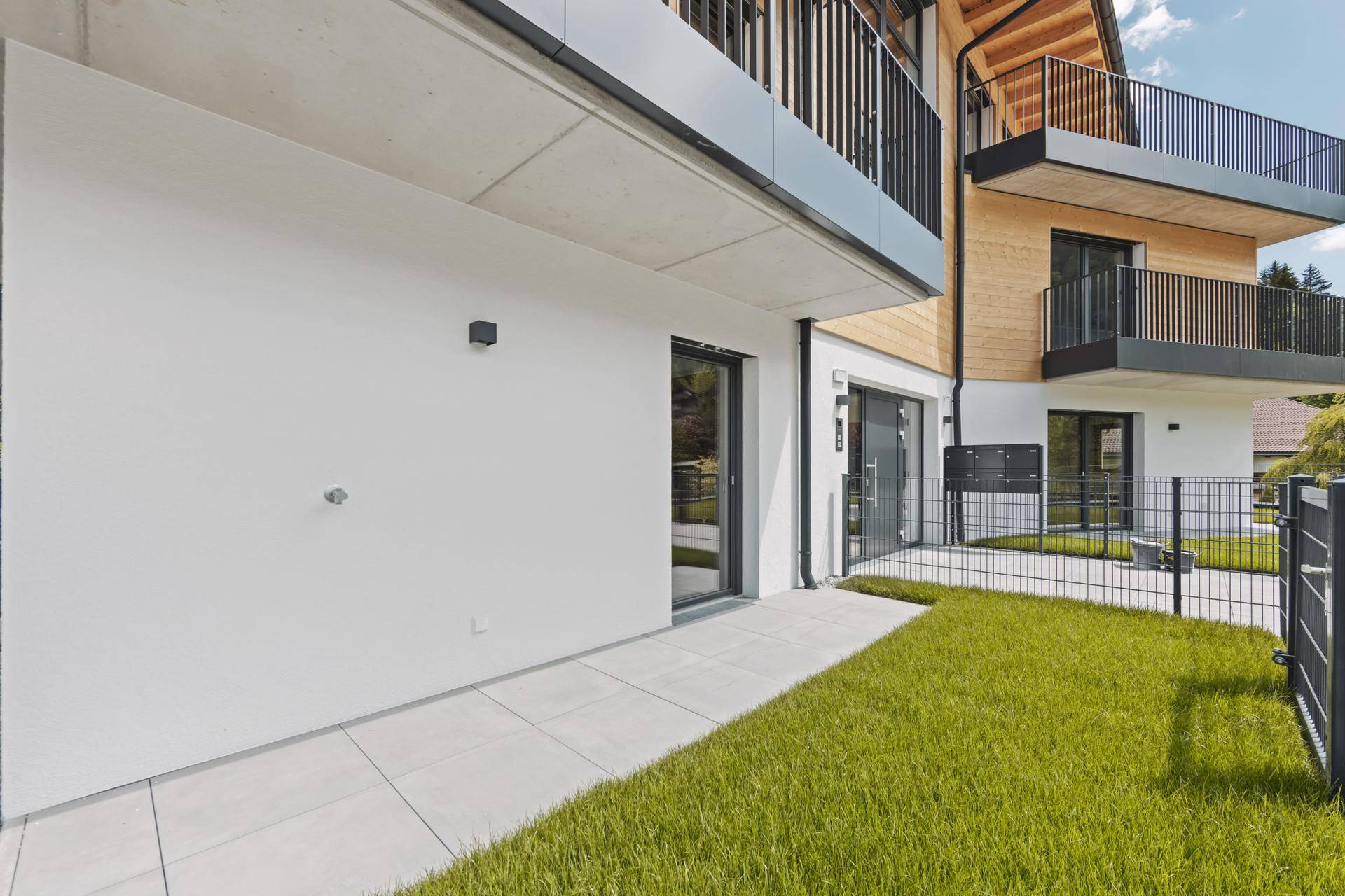 Gepflegte Grünfläche und gepflasterte Terrasse vor einem modernen Wohngebäude mit Balkonen.