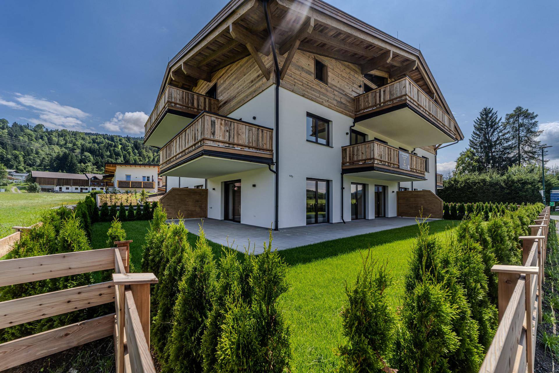 Modernes Gebäude mit Holzfassade und Balkonen, umgeben von einem gepflegten Garten und grüner Landschaft.
