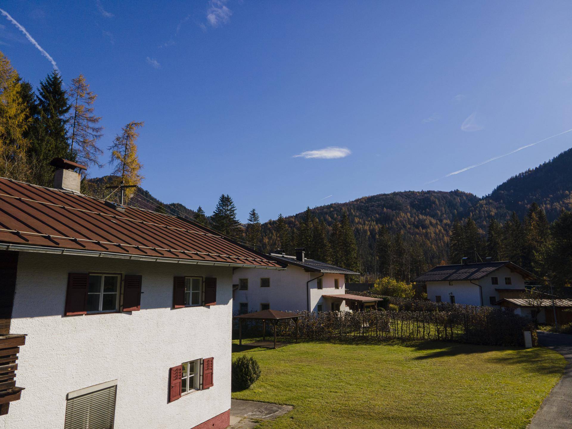 Seitenansicht des Hauses mit rotem Dach und Fensterläden, umgeben von grünen Wiesen und Bergen unter blauem Himmel.