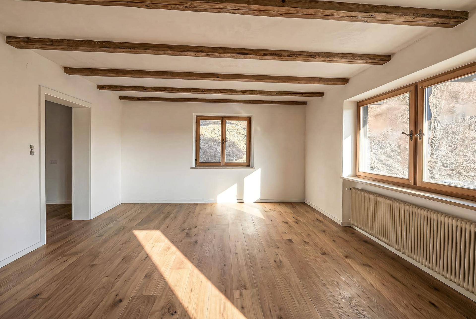 Heller Raum mit Holzbalkendecke, Dielenboden und großen Fenstern, die viel Licht hereinlassen.