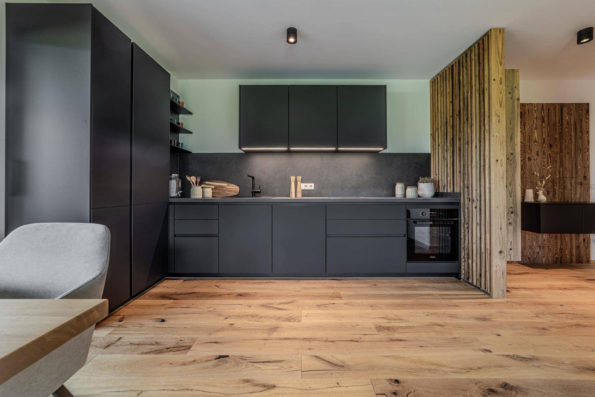 Moderne Küche mit dunklen Schränken, Holzboden und einer stilvollen Holztrennwand im offenen Wohnkonzept.