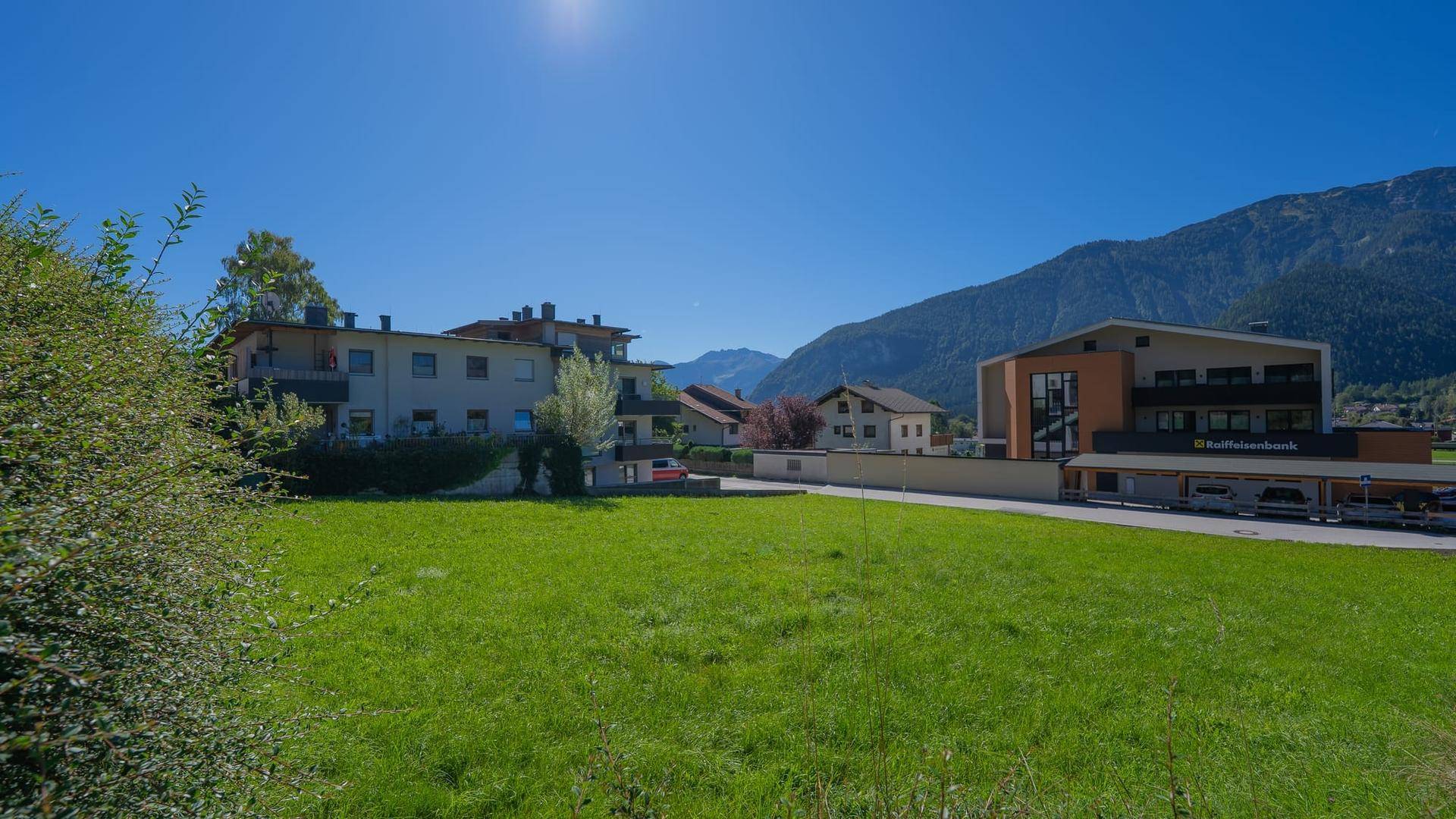 Sonnige Landschaft mit Gebäuden und Bergen im Hintergrund, umgeben von grünen Wiesen und Bäumen.