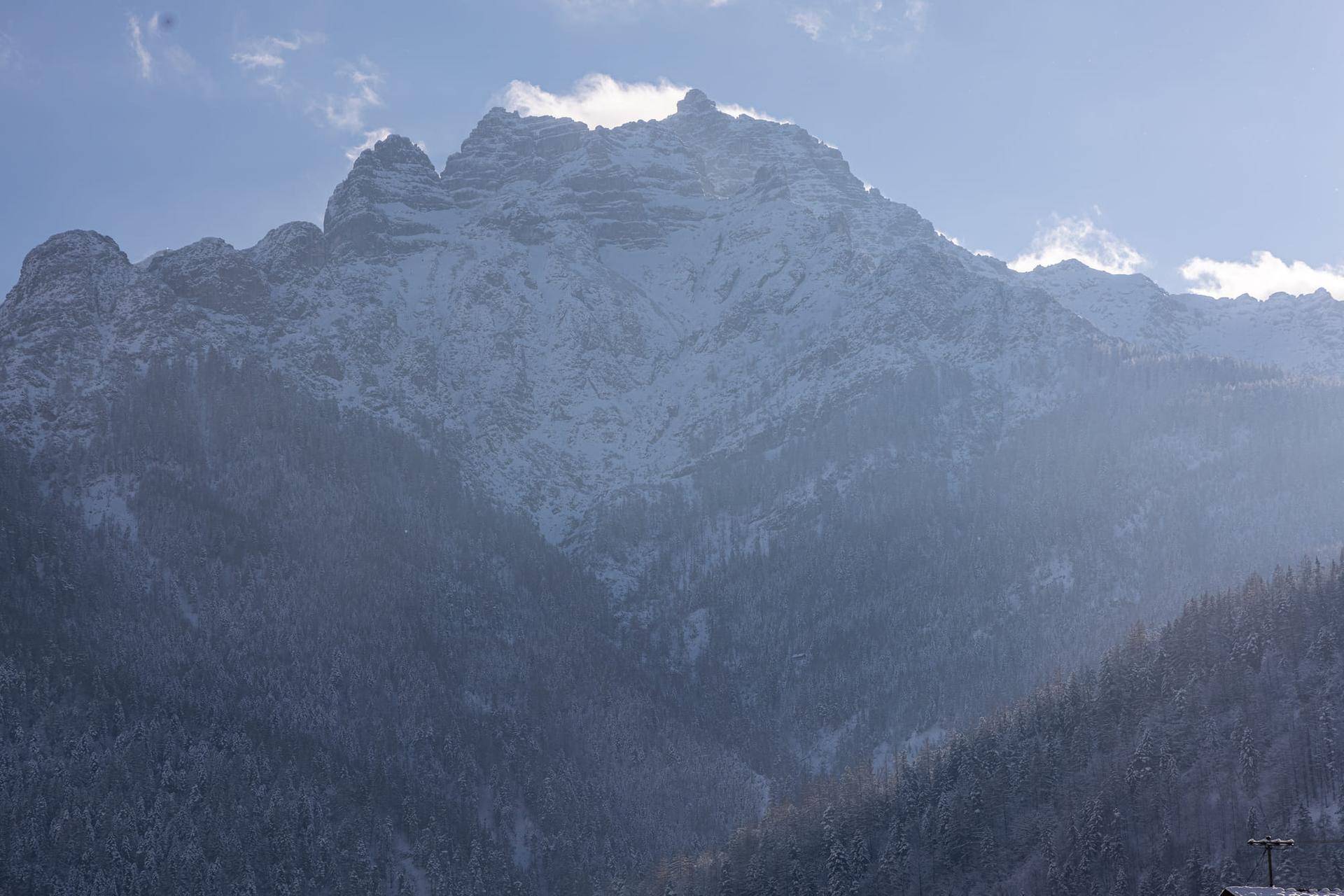 Nahaufnahme eines schneebedeckten Berggipfels mit Wolkenfetzen unter einem hellen Winterhimmel.