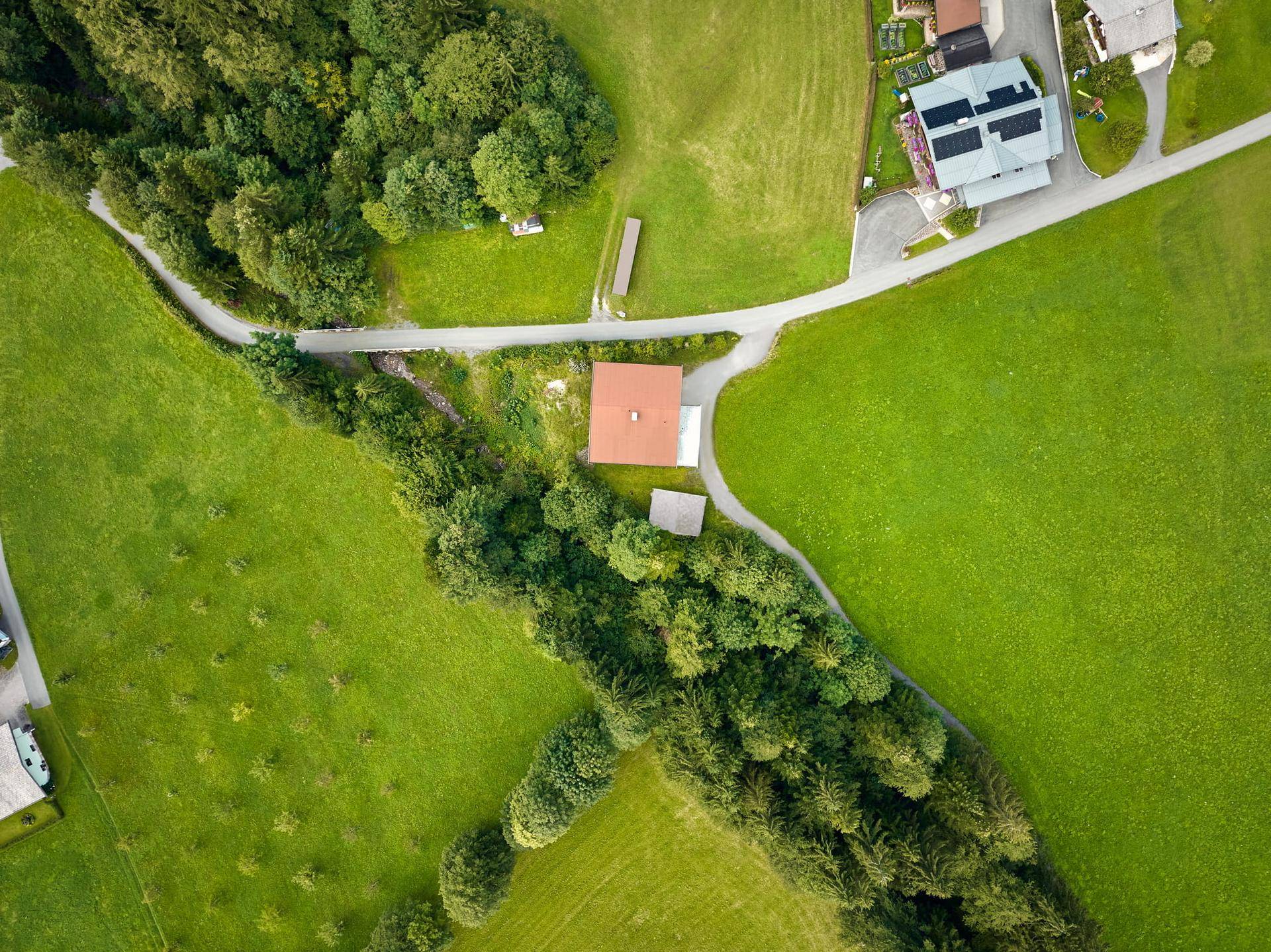 Luftaufnahme eines Hauses mit rotem Dach, umgeben von grünen Wiesen und Bäumen, mit einer kurvigen Straße.