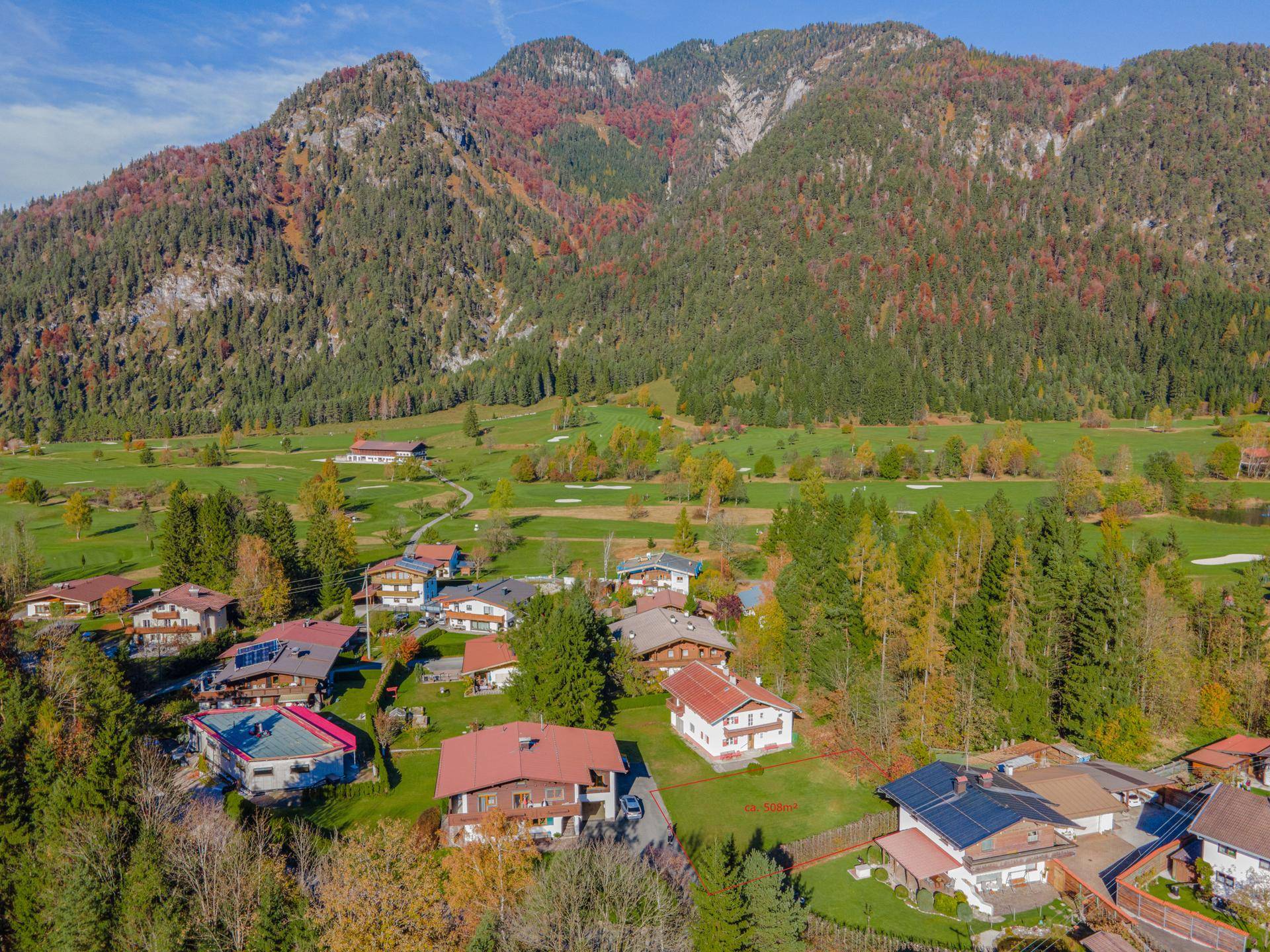 Luftaufnahme eines malerischen Dorfes in einem Tal, umgeben von bewaldeten Bergen und Golfplätzen.