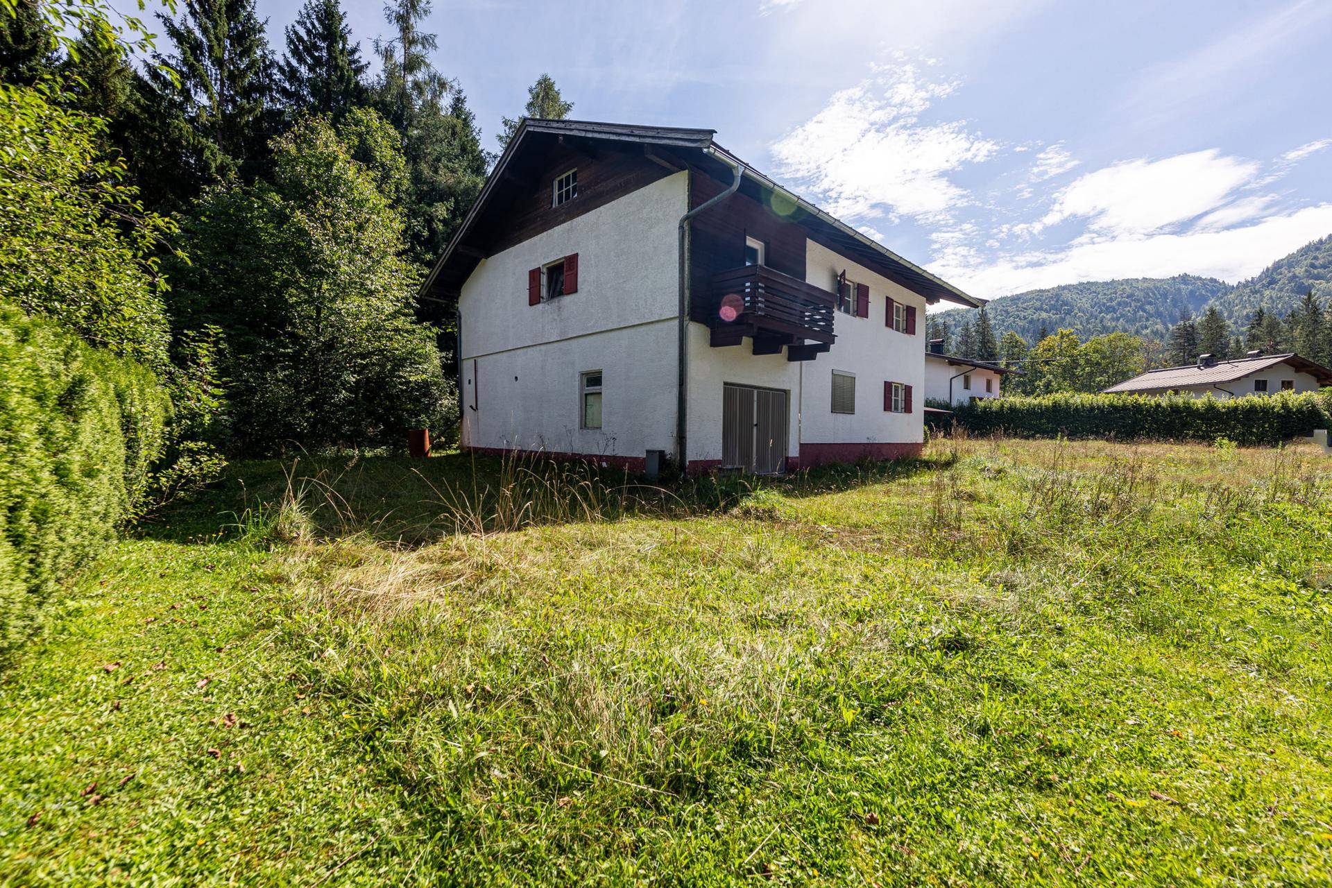 Hausansicht von der Seite mit kleinem Balkon und umgebender Natur, im Hintergrund sind Berge sichtbar.