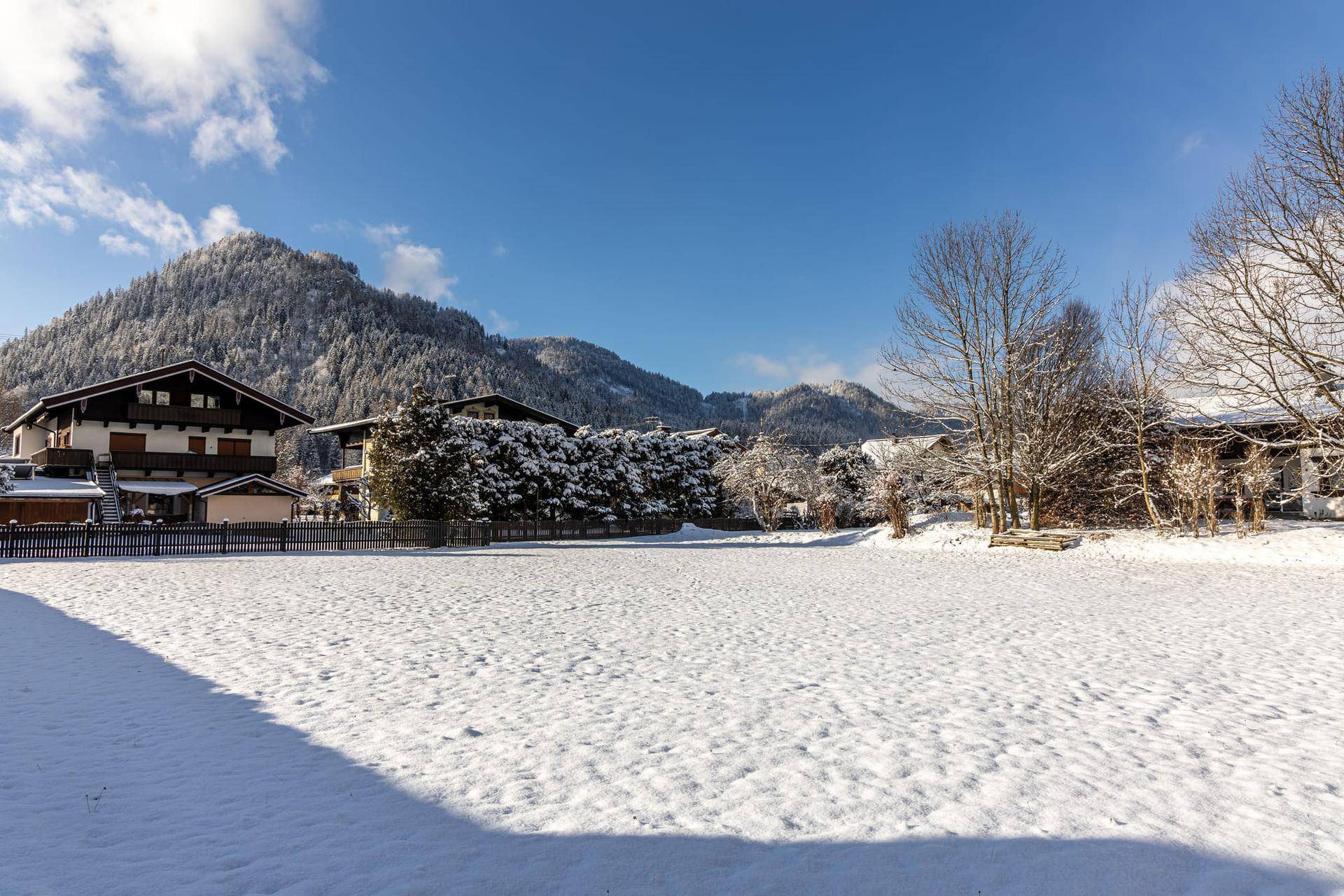 Breite schneebedeckte Fläche mit umliegenden Häusern und bewaldeten Bergen unter klarem blauem Himmel.