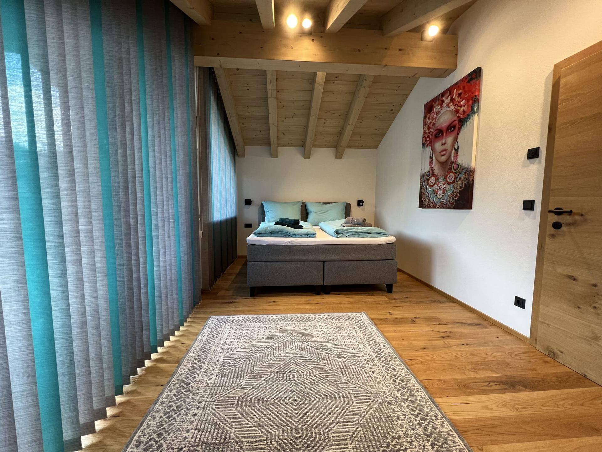 Gemütliches Schlafzimmer mit Holzbalkendecke, Doppelbett und großem Fenster mit Jalousien.