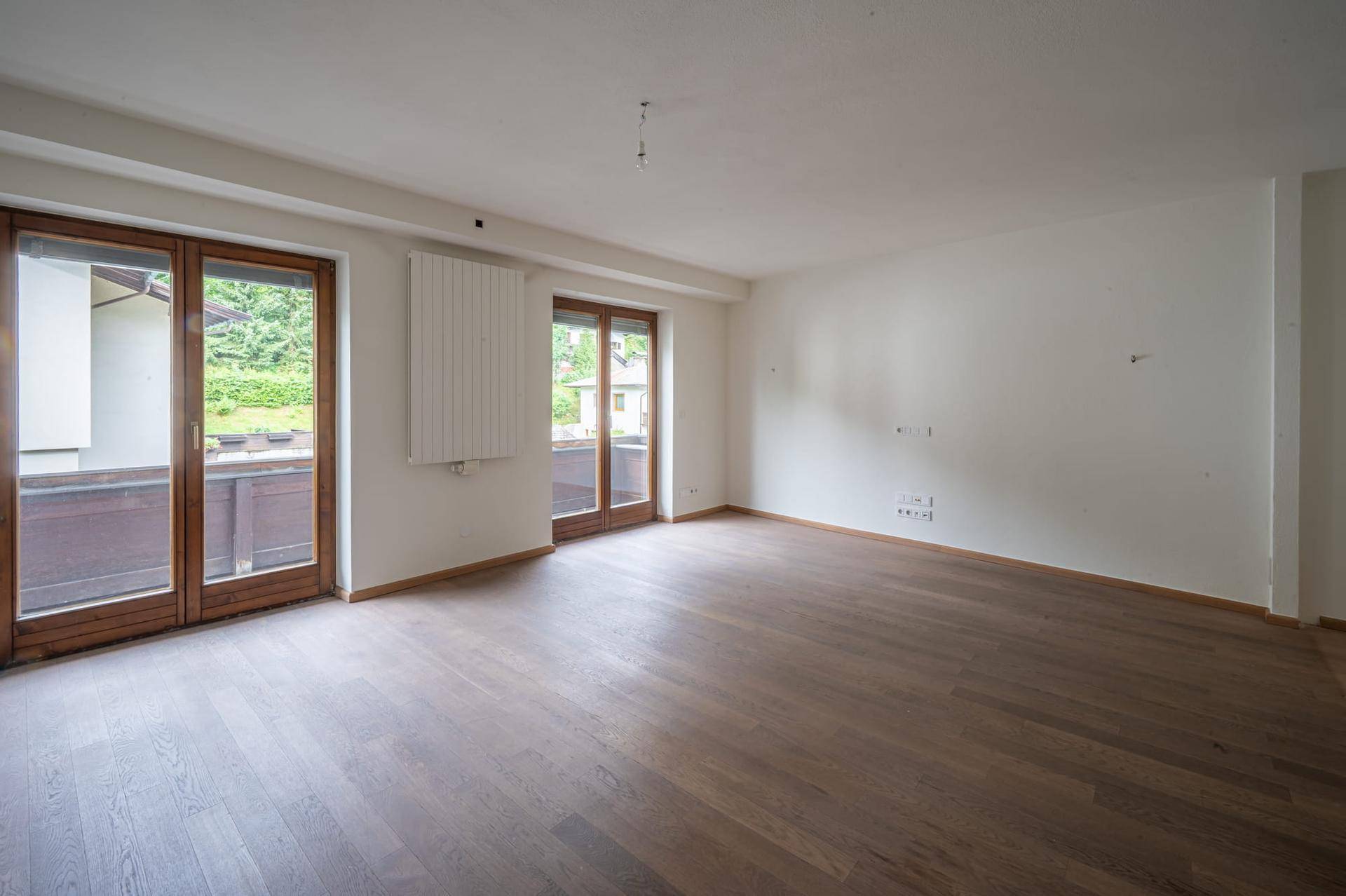 Leeres Zimmer mit dunklem Holzboden, weißen Wänden und zwei großen Glastüren, die zu einem Balkon führen.