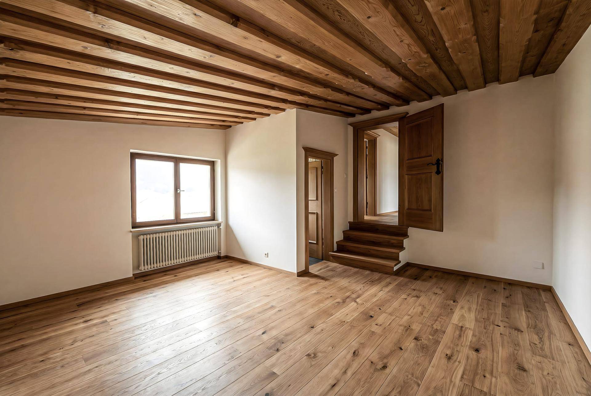 Lichtdurchfluteter Raum mit Holzbalkendecke, Dielenboden und Zugang zu weiteren Zimmern.
