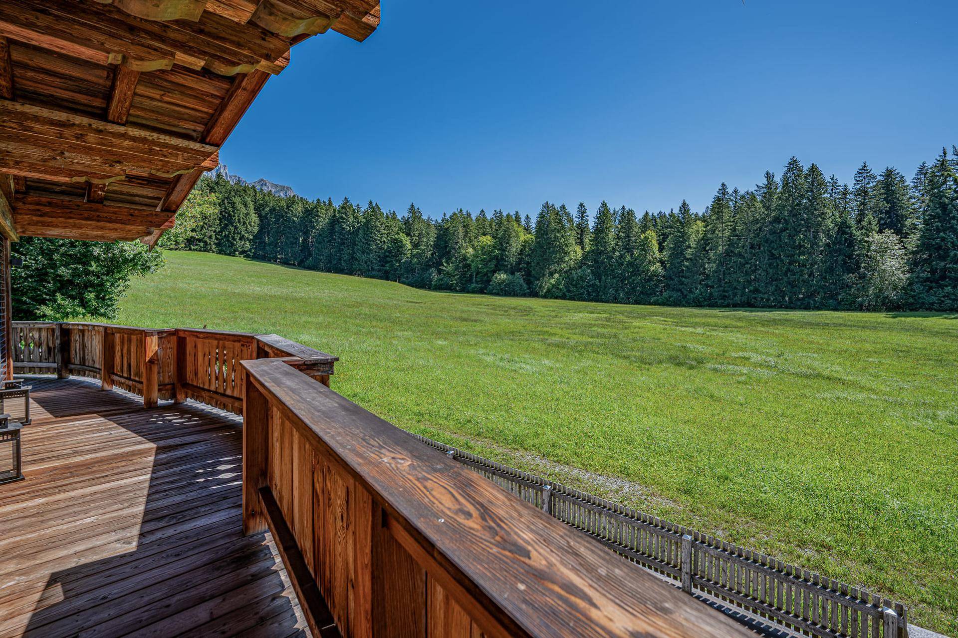 Geräumiger Holzbalkon mit Blick auf weite grüne Wiesen und dichte Wälder unter blauem Himmel.