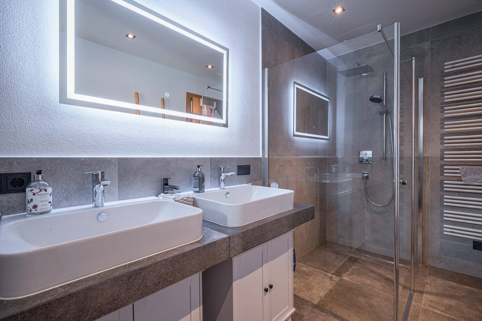 Modernes Badezimmer mit Doppelwaschbecken, beleuchteten Spiegeln und einer ebenerdigen Dusche.
