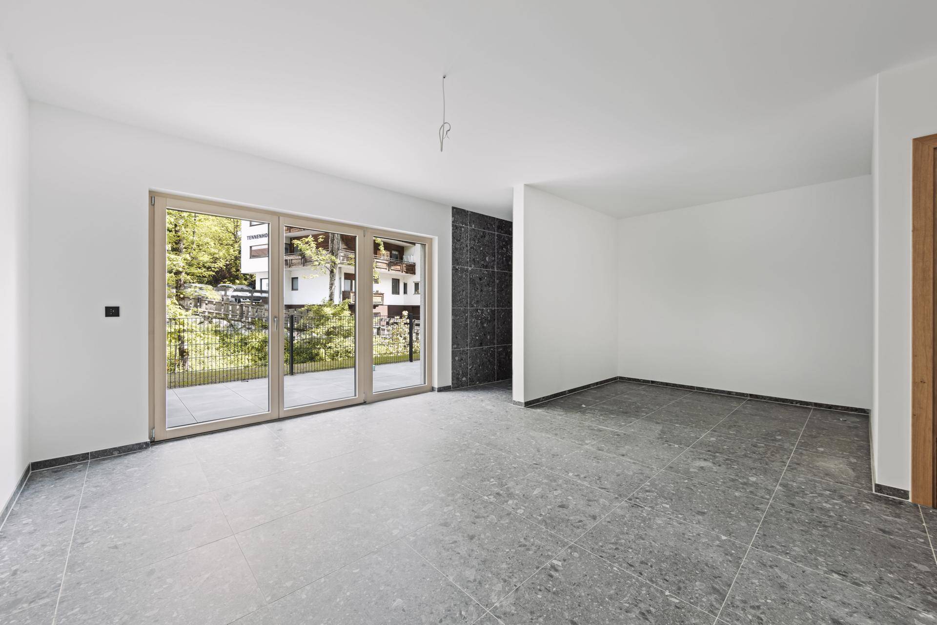 Geräumiger, unmöblierter Raum mit dunklen Fliesen und breiter Fensterfront zum Balkon.