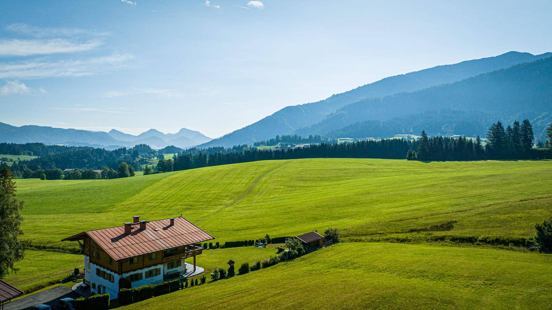 Panoramablick auf das charmante Landhaus inmitten weitläufiger grüner Felder und majestätischer Berge.
