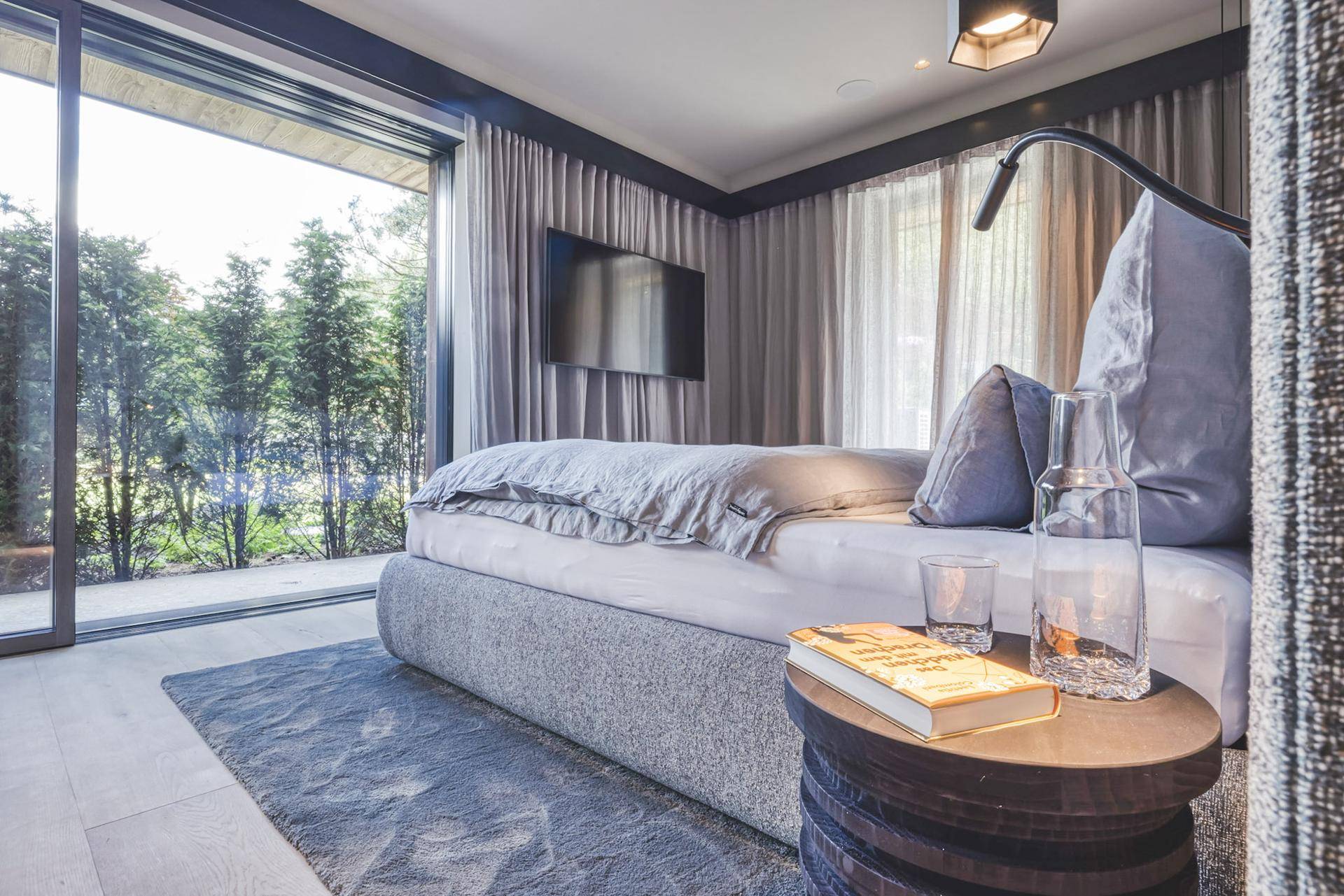 Gemütliches Schlafzimmer mit großem Bett, Wandfernseher und raumhohen Fenstern mit Blick ins Grüne.