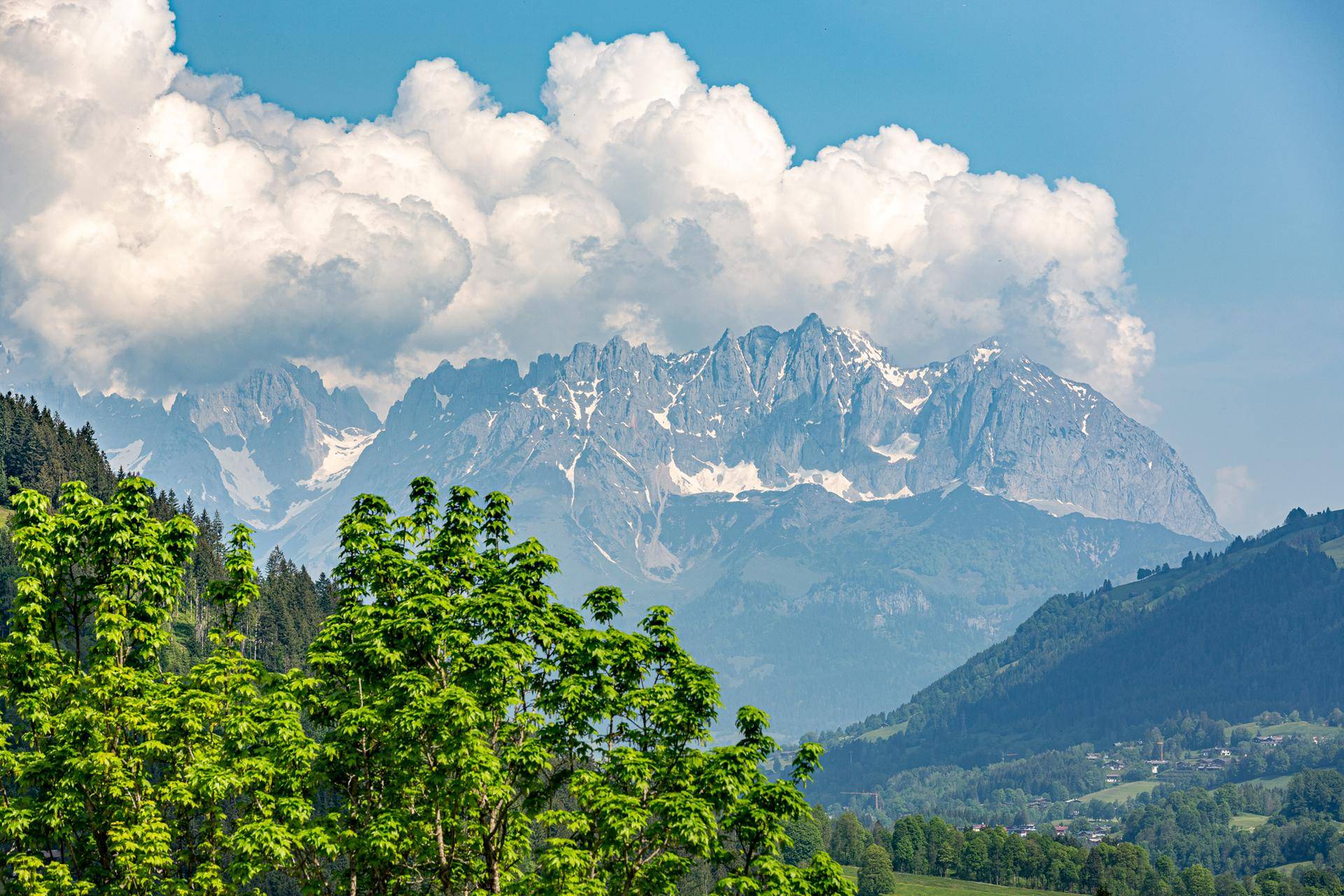 Malerische Berglandschaft mit grünen Hügeln und schneebedeckten Gipfeln unter blauem Himmel.