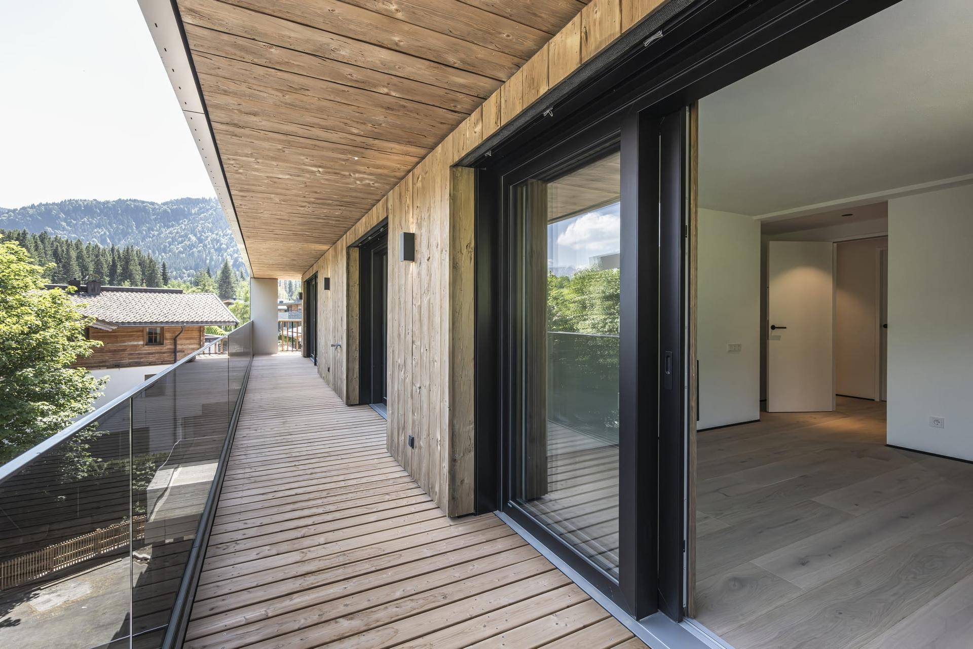 Langer Holzbalkon mit Glasgeländer und Bergblick, zugänglich von mehreren Räumen.