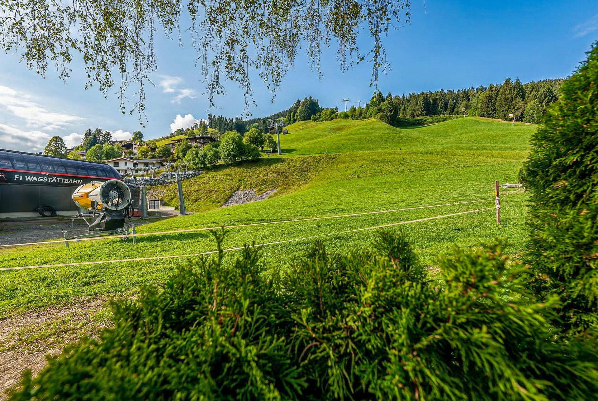 Grüne Skipiste mit Gondelbahn und Beschneiungsanlage unter strahlend blauem Himmel.