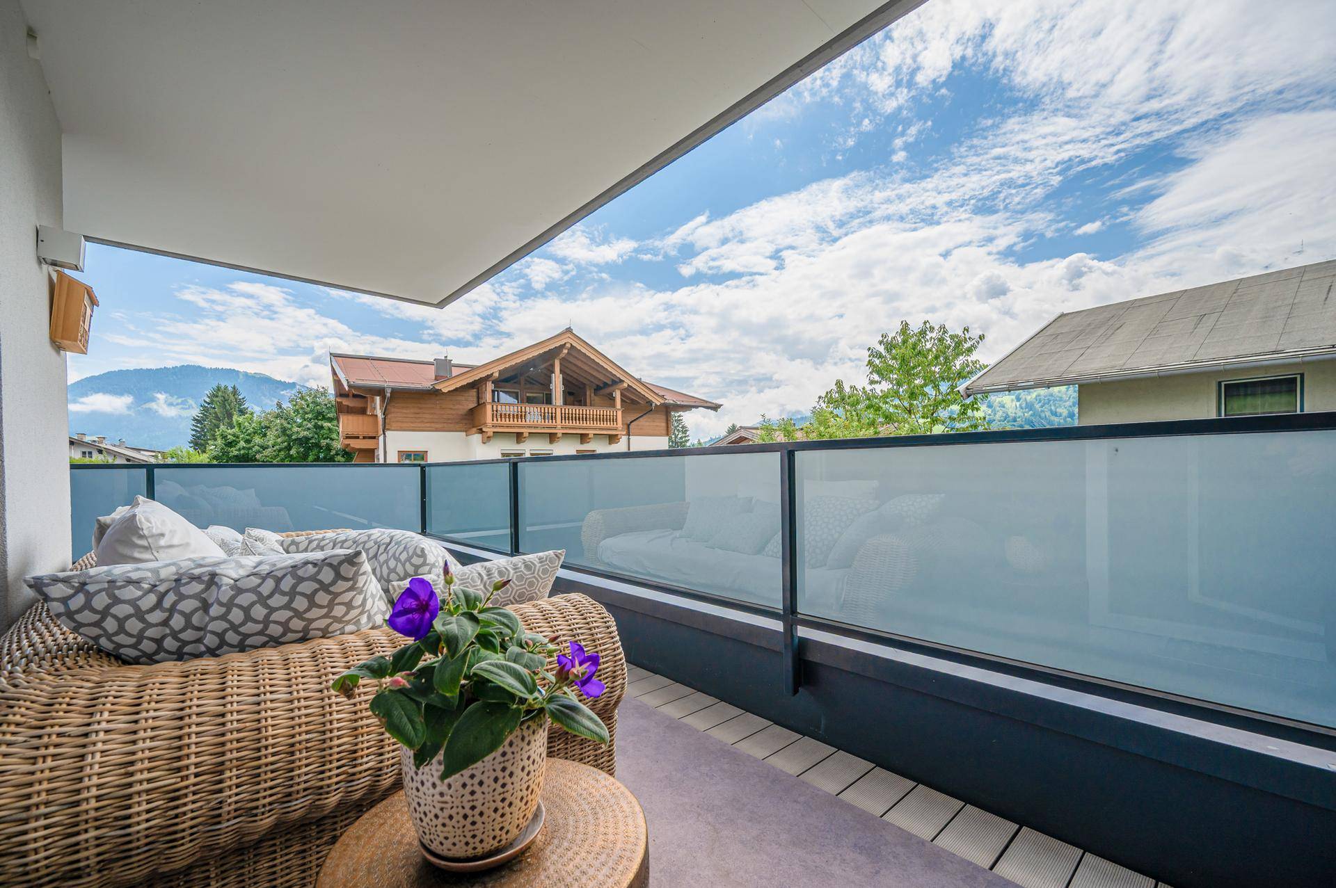 Gemütlicher Balkon mit Rattanmöbeln, Glasgeländer und schönem Blick auf die umliegende Berglandschaft.