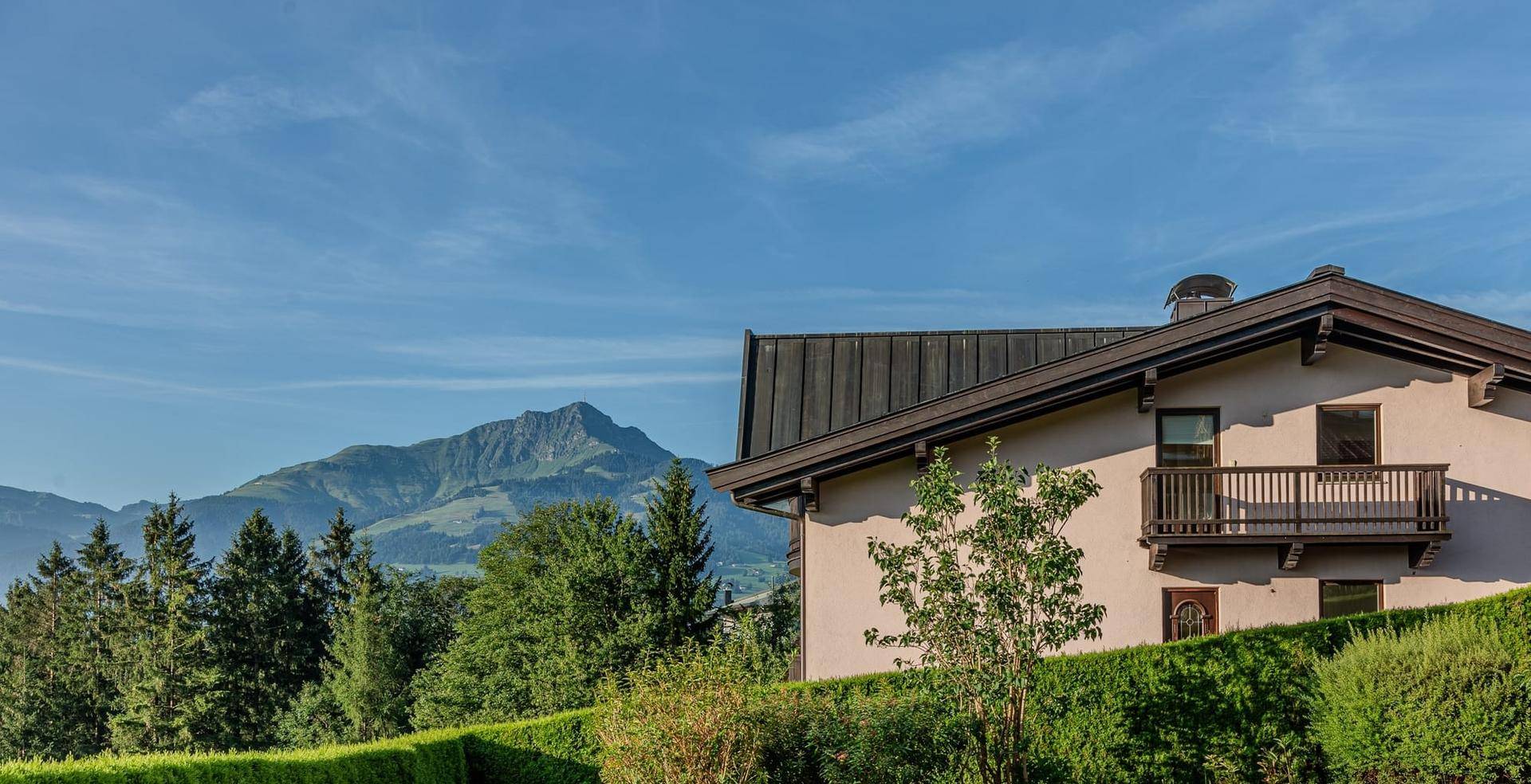 Traditionelles Haus mit Holzbalkon und Bergblick, umgeben von einer grünen Hecke.