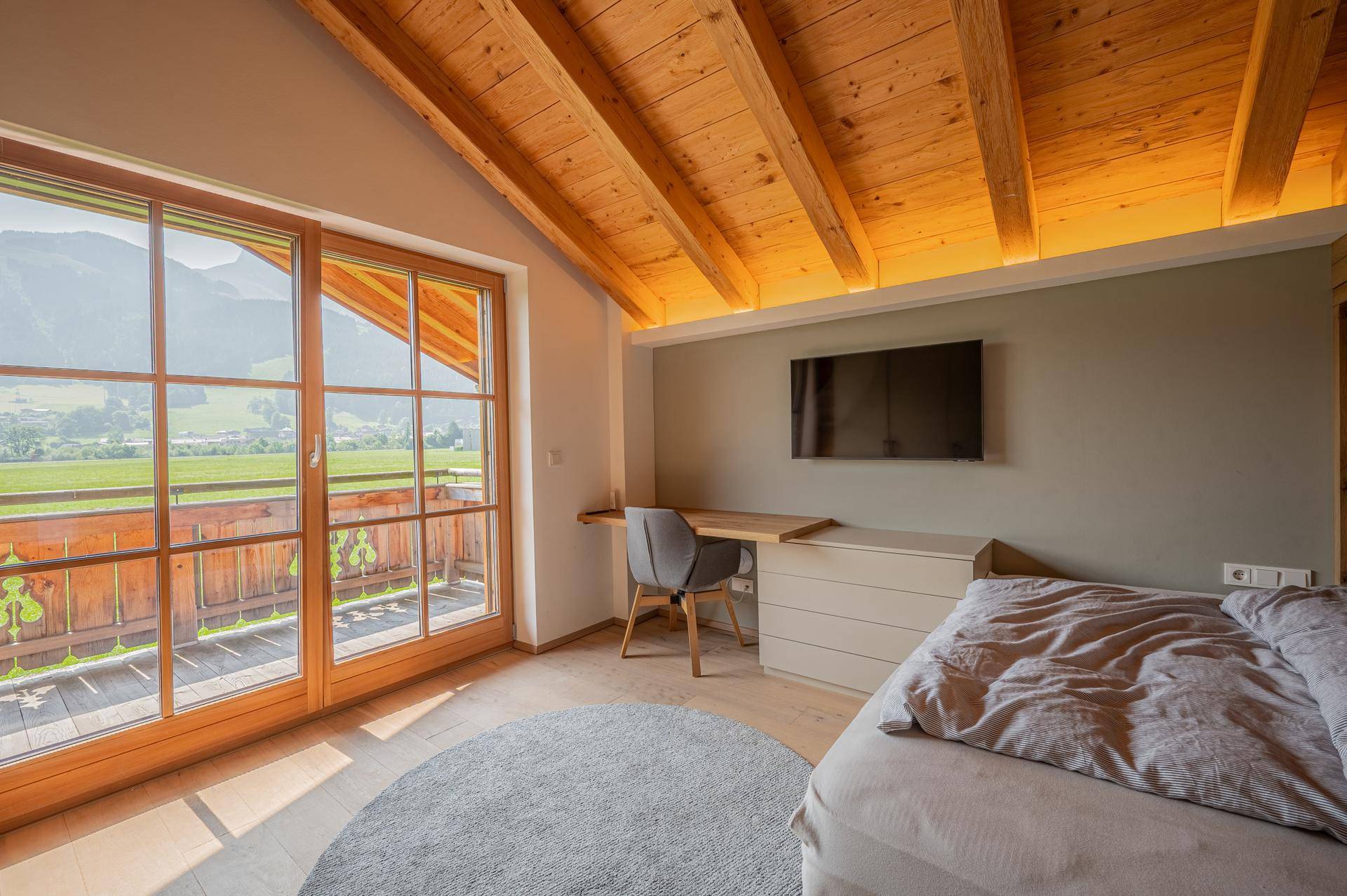 Helles Schlafzimmer mit Holzbalkendecke, Schreibtisch und Zugang zu einem Balkon mit Bergblick.