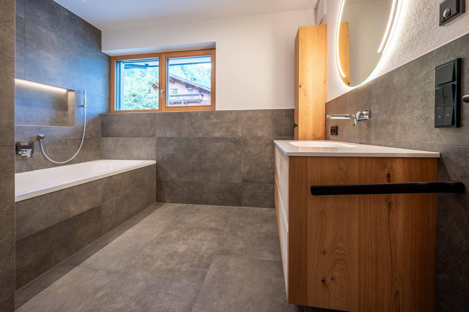 Modernes Badezimmer mit Badewanne, Holzwaschtisch und großen grauen Fliesen.