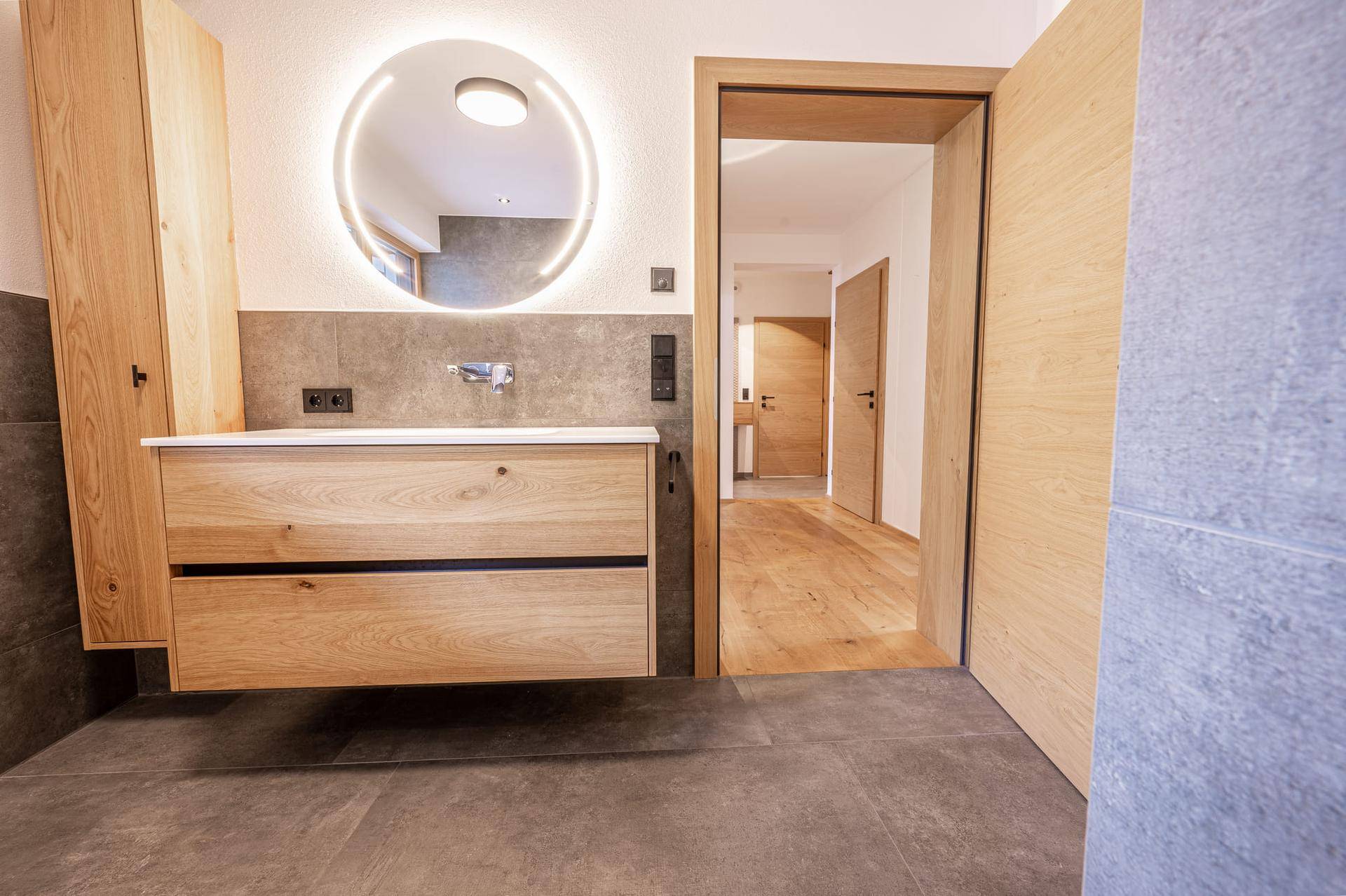 Elegantes Badezimmer mit schwebendem Holzwaschtisch, rundem beleuchtetem Spiegel und Zugang zum Flur.
