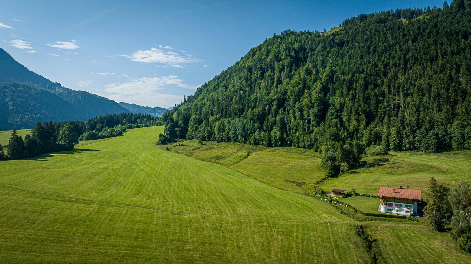 Weitläufige Landschaft mit einem traditionellen Haus, umgeben von grünen Hügeln, Wäldern und Bergen.
