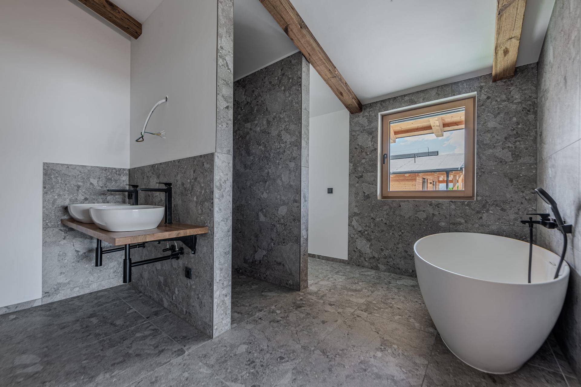 Modernes Badezimmer mit freistehender Badewanne, Doppelwaschbecken und grauen Fliesen, unter sichtbaren Holzbalken.