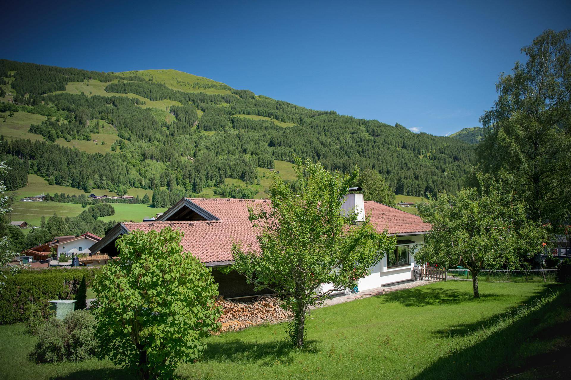 Idyllisches Haus mit rotem Ziegeldach, umgeben von grünen Wiesen und bewaldeten Bergen.