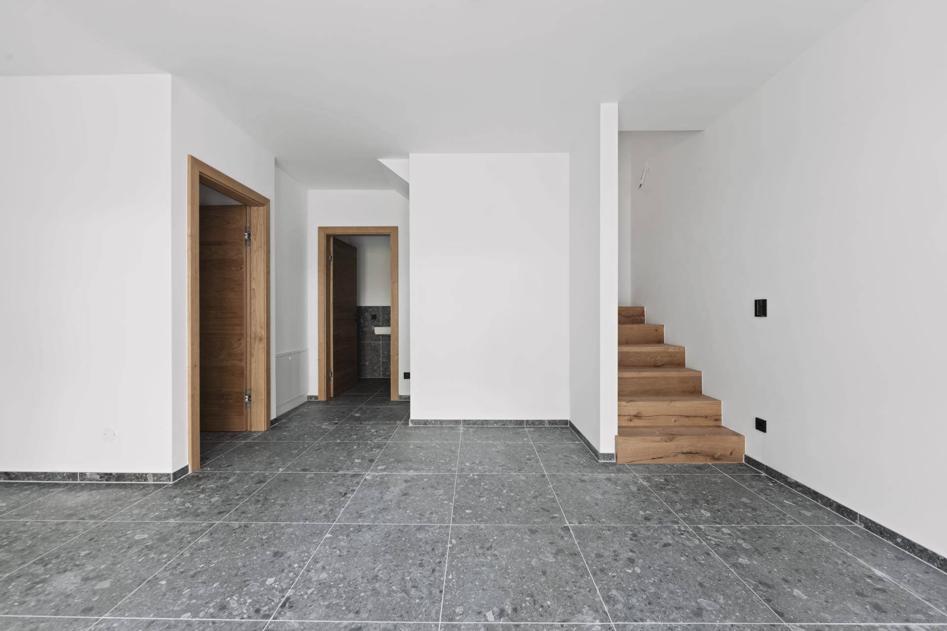 Eingangsbereich mit moderner Holztreppe und dunklem Fliesenboden, führt zu weiteren Räumen.