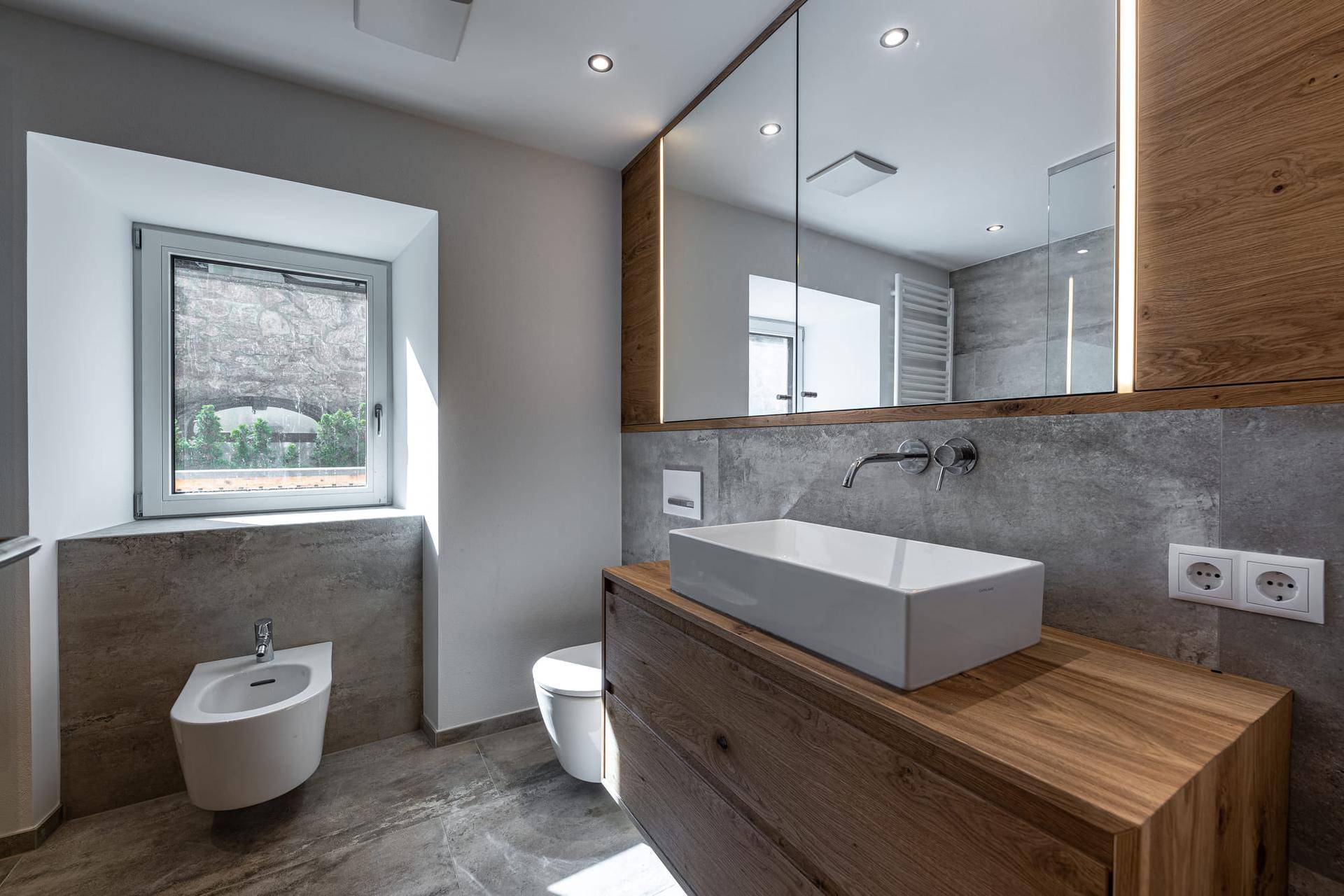 Modernes Badezimmer mit Holzelementen, großem Waschbecken, Bidet und Fenster für natürliches Licht.