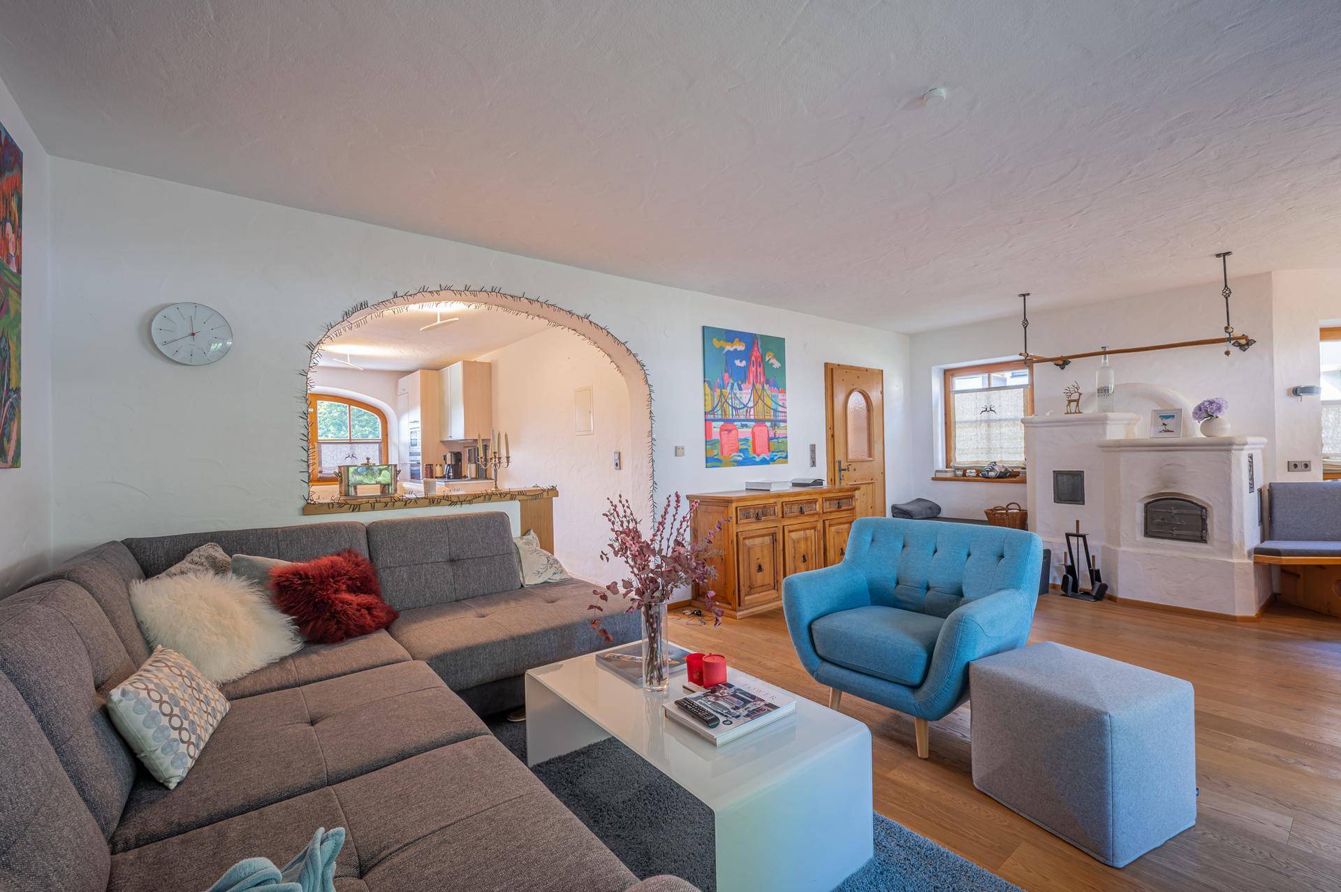 Helles Wohnzimmer mit großem Sofa, Kachelofen und offenem Durchgang zur Küche.