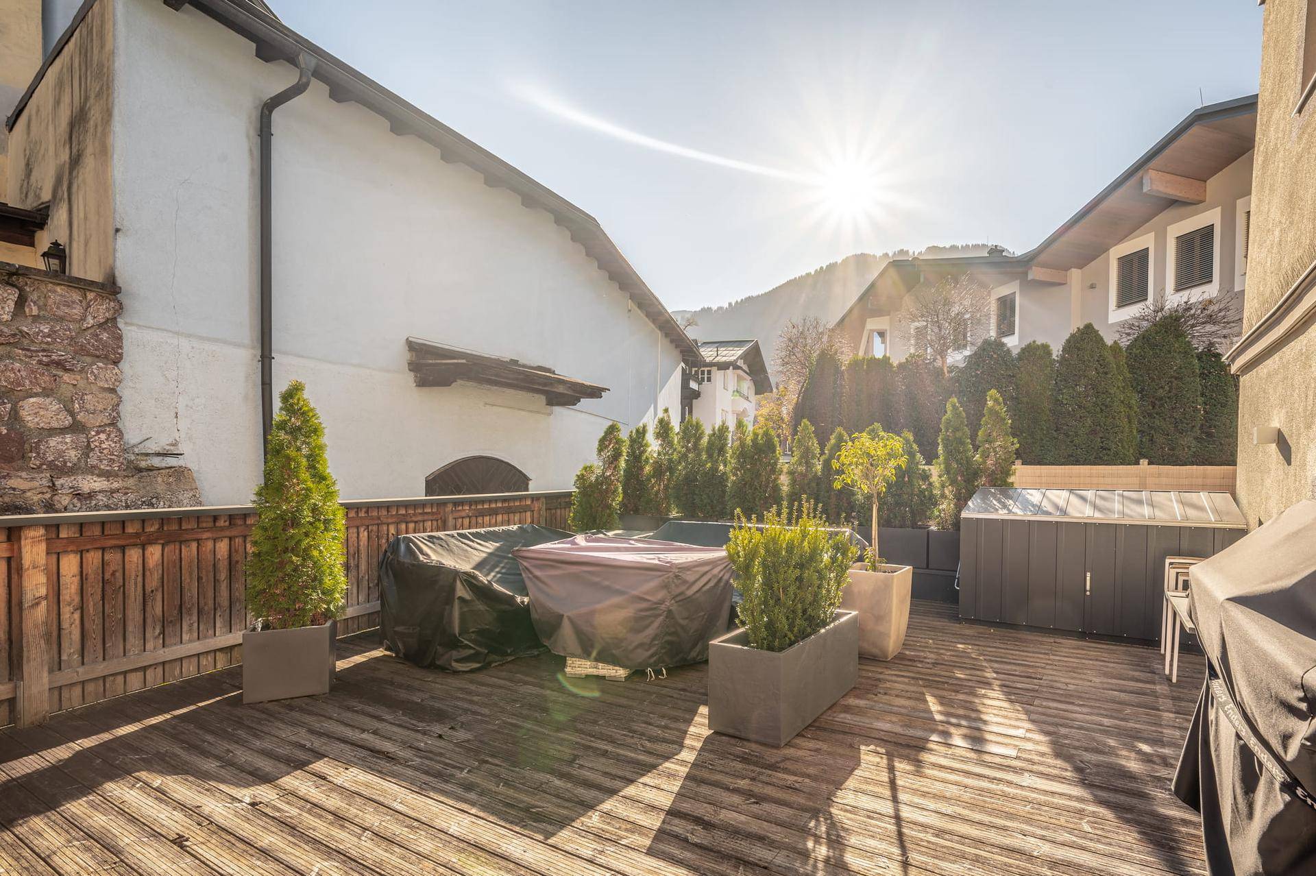 Sonnige Holzterrasse mit Gartenmöbeln und Blick auf die umliegenden Berge, umgeben von Grünpflanzen.