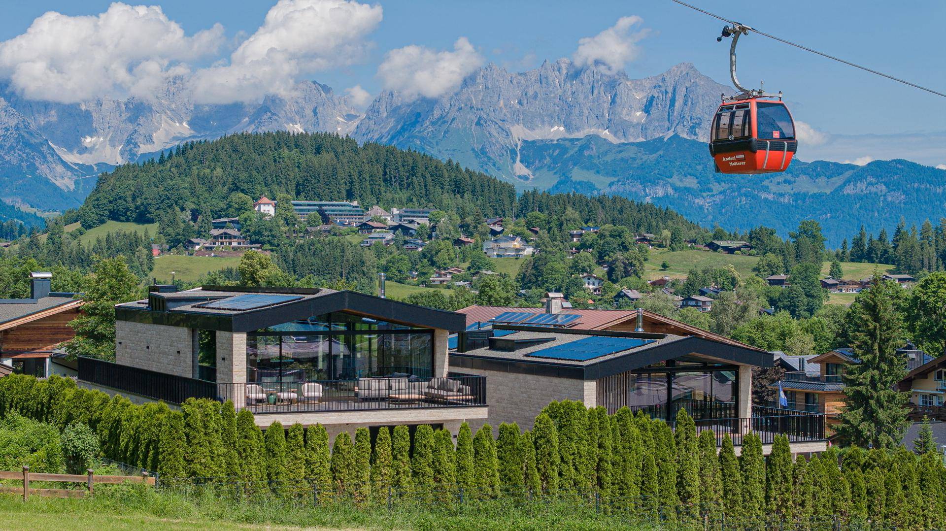 Moderne Villen mit Flachdach und großen Glasfronten vor alpiner Berglandschaft mit Seilbahn.