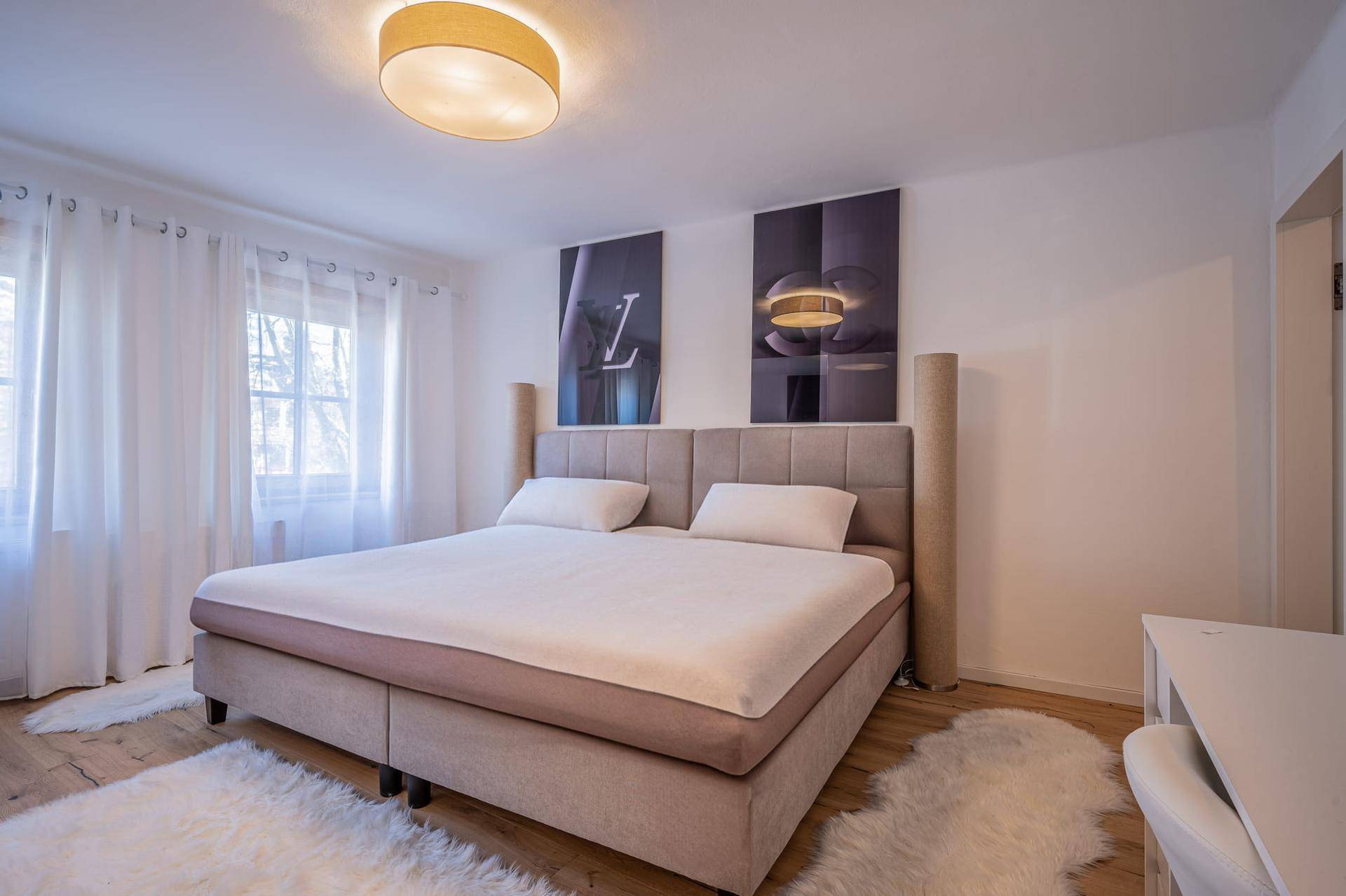 Schlafzimmer mit großem Doppelbett, Holzboden und hellen Vorhängen an den Fenstern für viel Tageslicht.