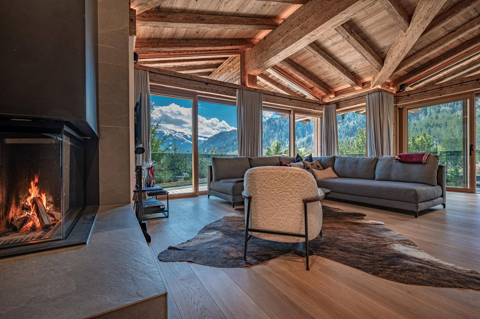 Gemütliches Wohnzimmer mit Kamin, Holzbalkendecke und Panoramafenstern mit Bergblick.