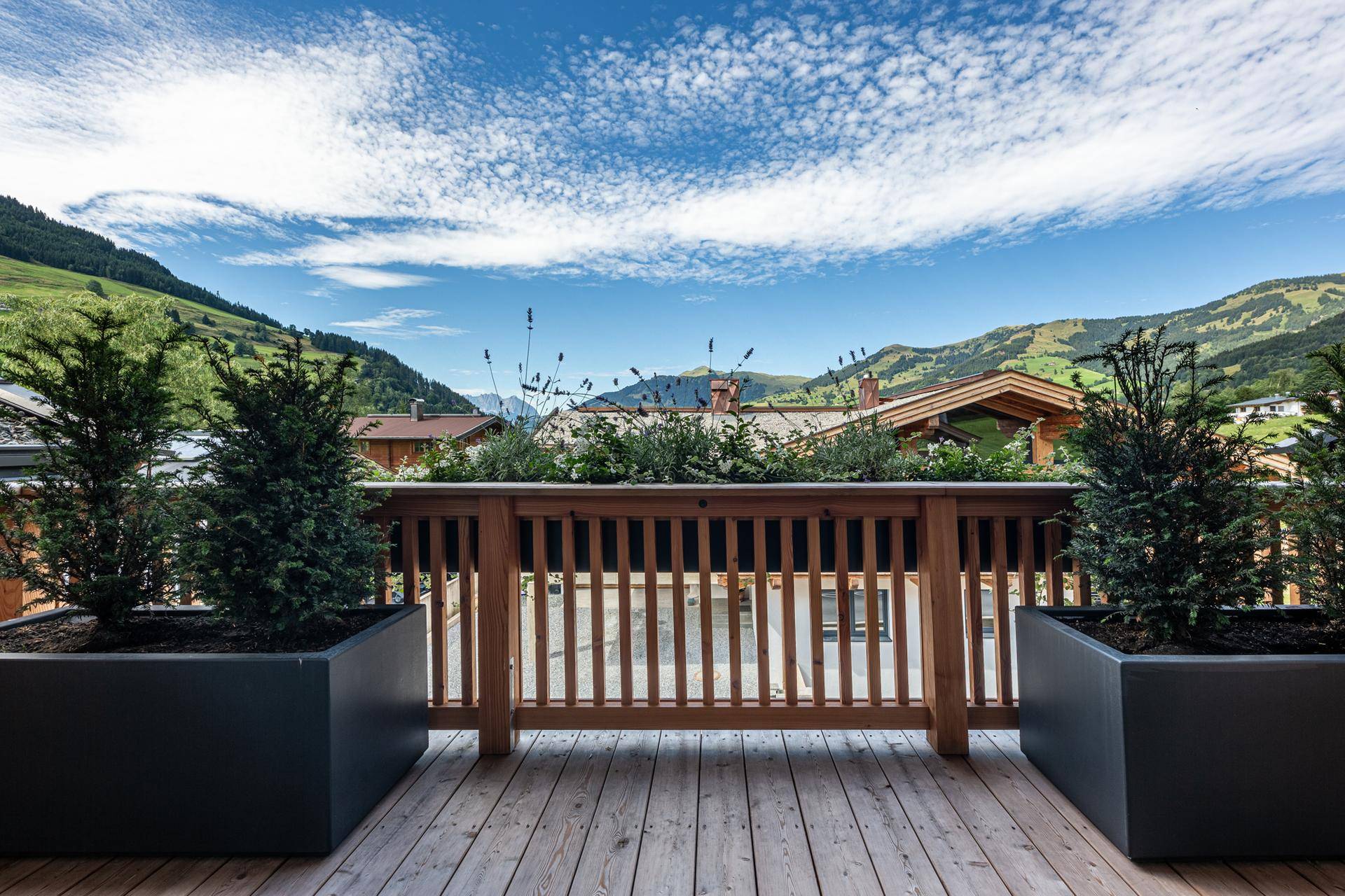 Geräumiger Holzbalkon mit Pflanzen und weitem Blick auf die alpine Landschaft und umliegende Häuser.