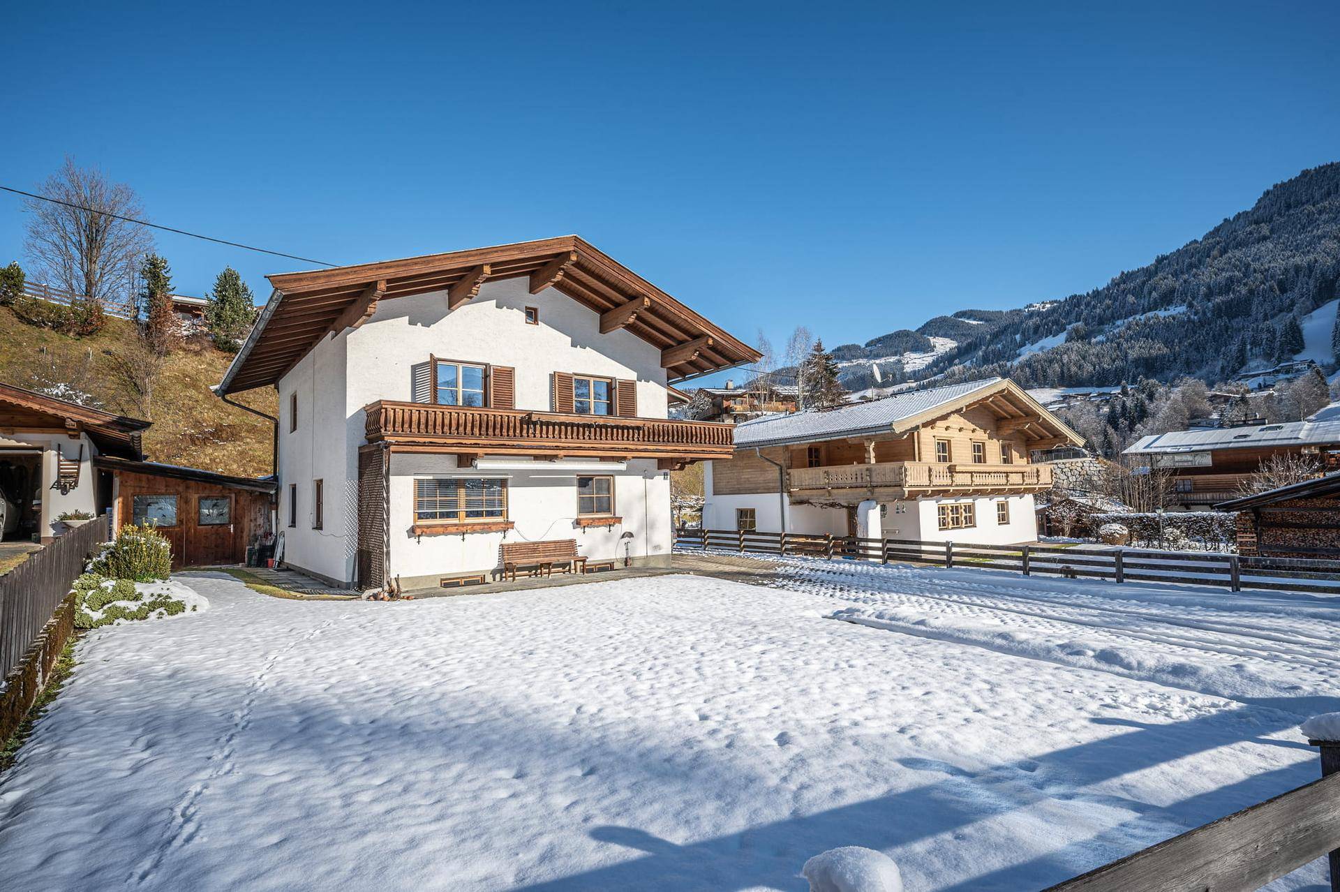 Traditionelles Haus mit Holzbalkonen und verschneitem Vorgarten vor einer Bergkulisse.