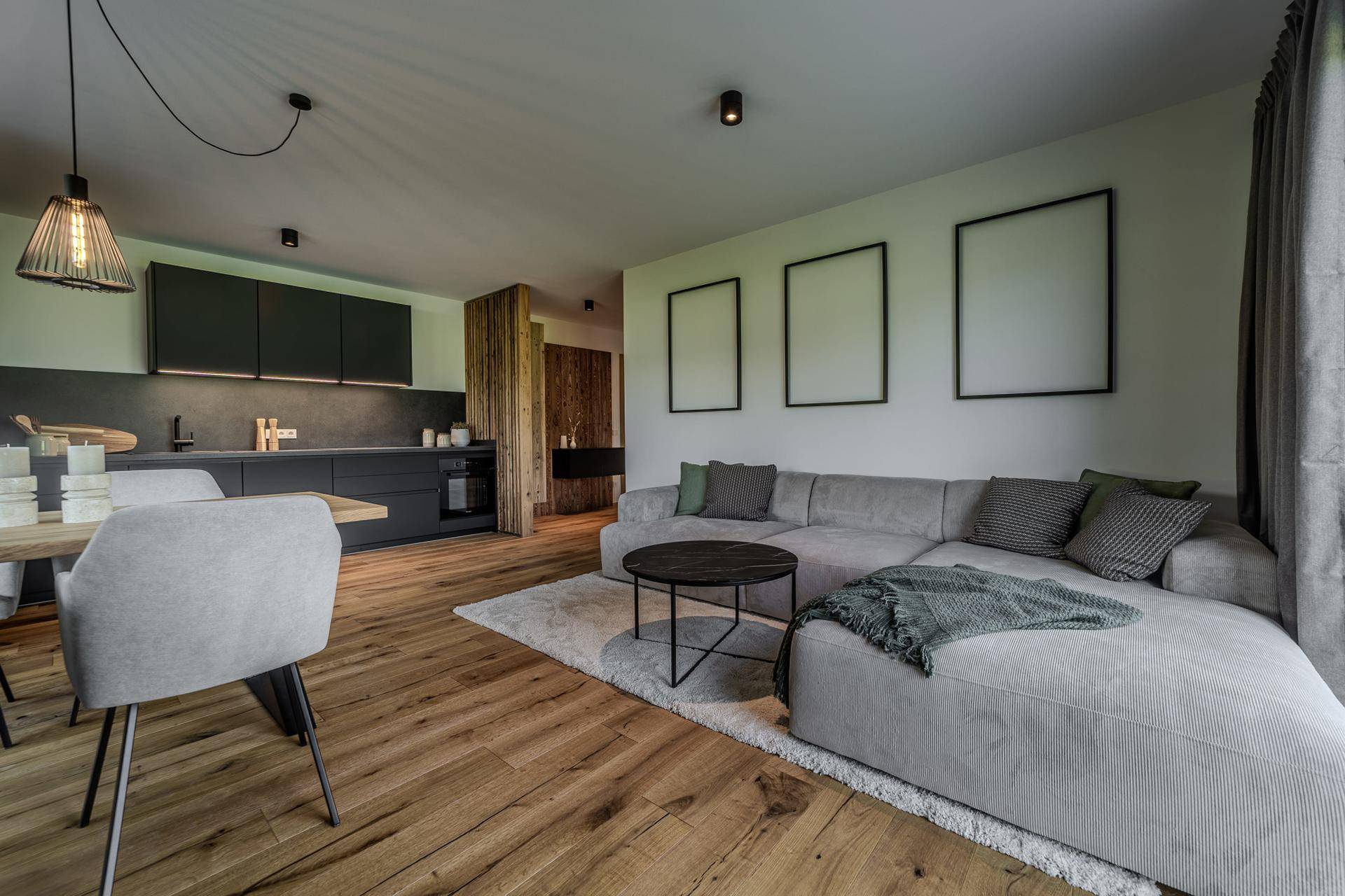 Offener Wohnbereich mit bequemer Couch, Holzboden und integrierter, moderner Küche in dunklen Farbtönen.