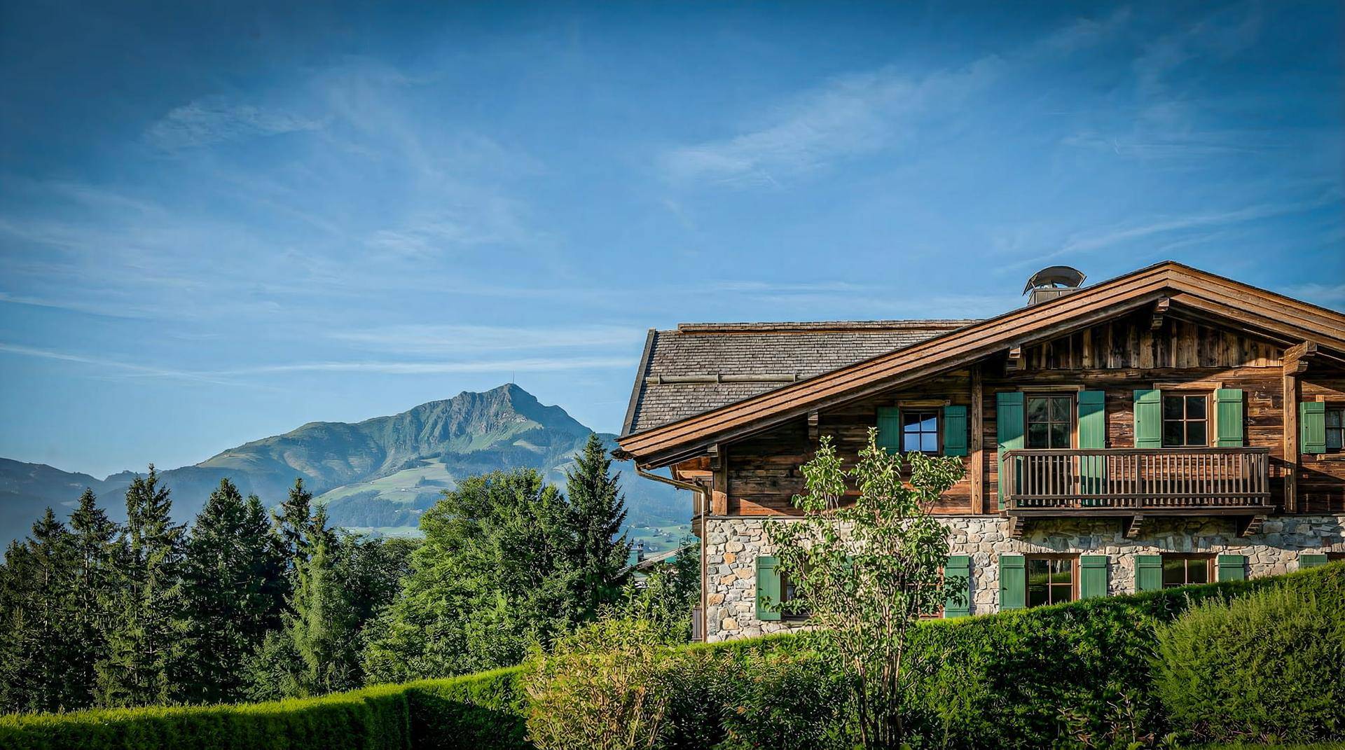 Traditionelles Holzchalet mit Steinfassade und grünen Fensterläden vor beeindruckender Bergkulisse.