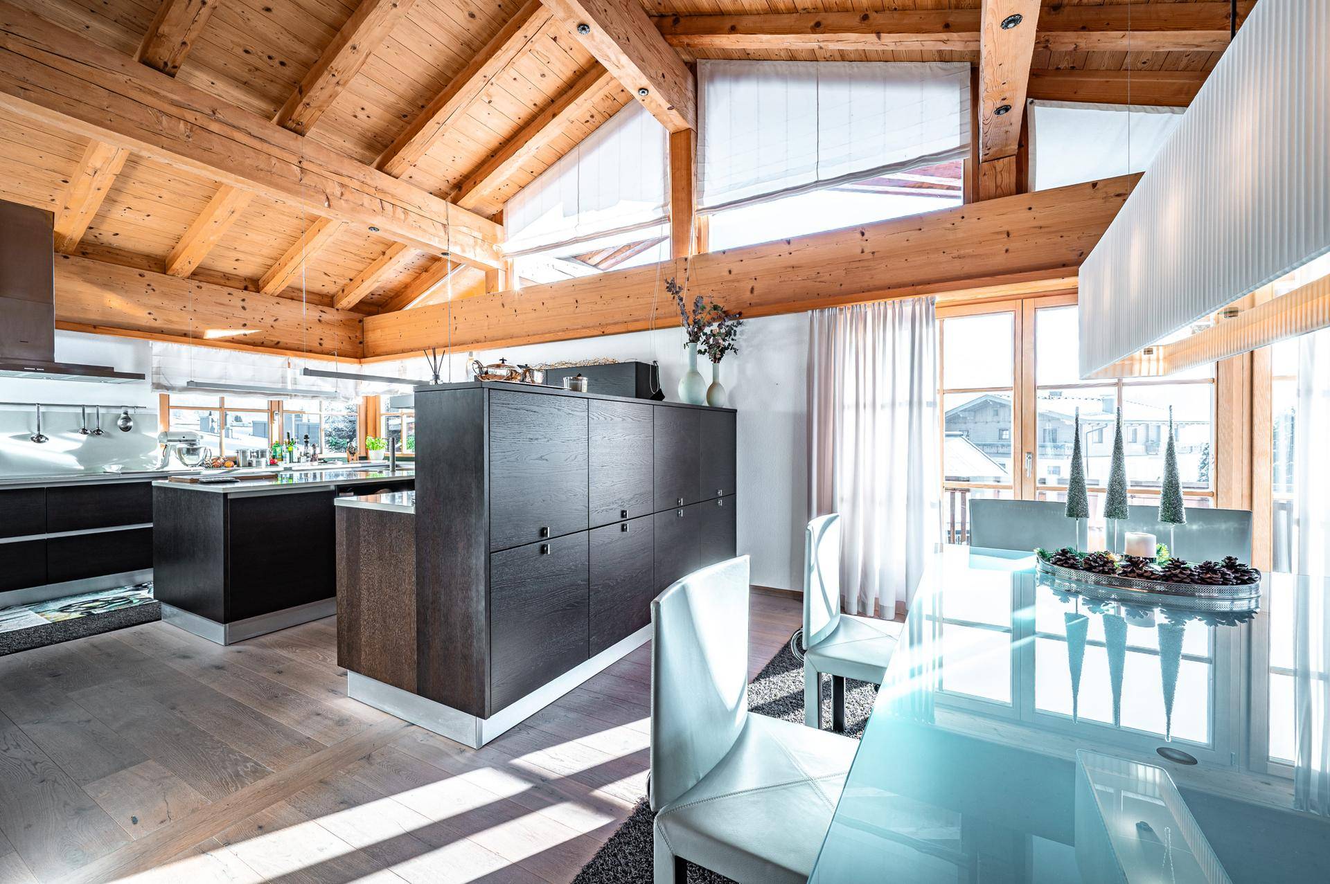 Moderne Küche mit dunklen Schränken, Holzbalkendecke und hellem Essbereich mit Glasstisch.