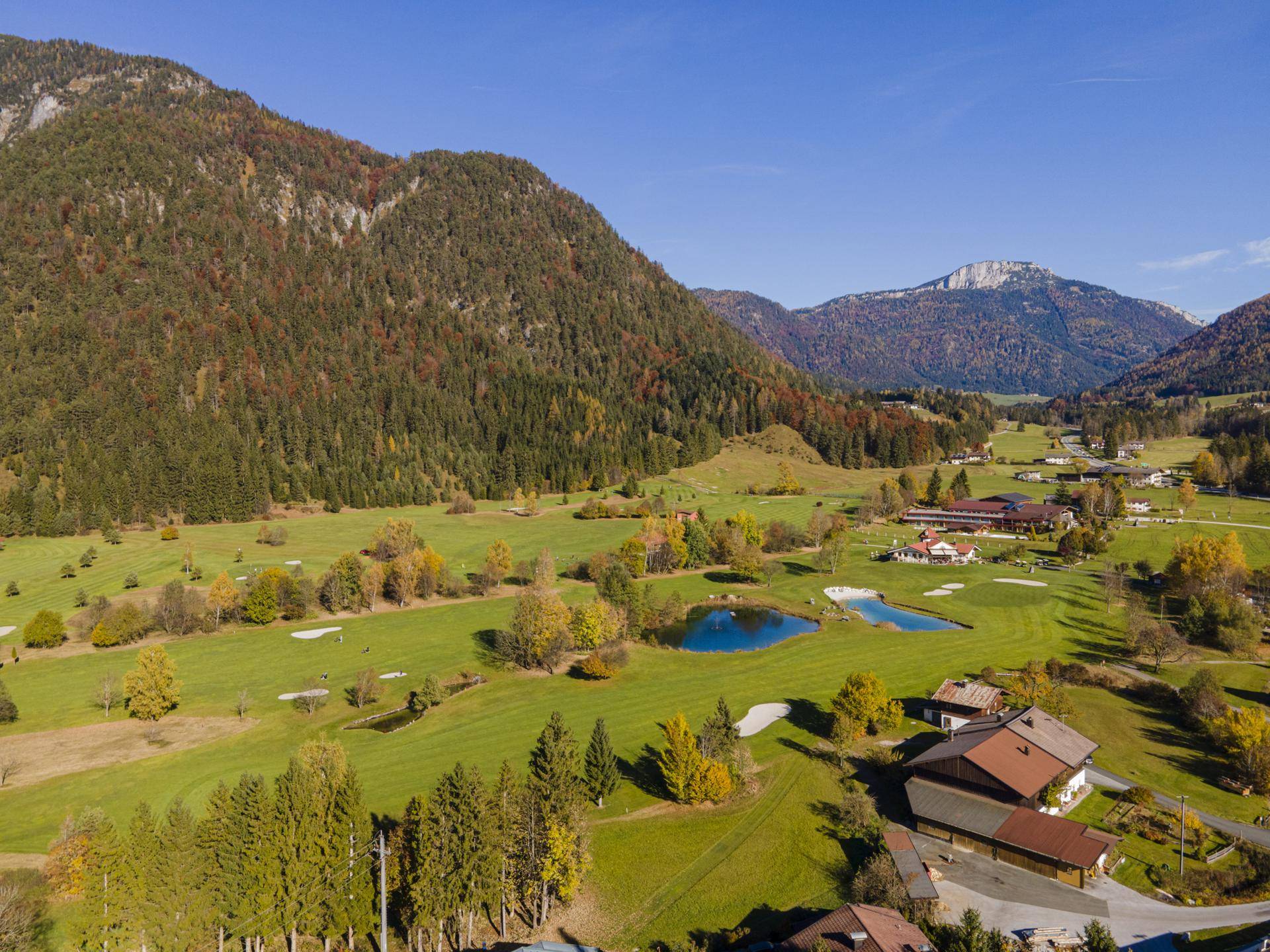 Weitläufige Luftaufnahme eines Tals mit Golfplatz, Teichen und Häusern, umgeben von bewaldeten Bergen.