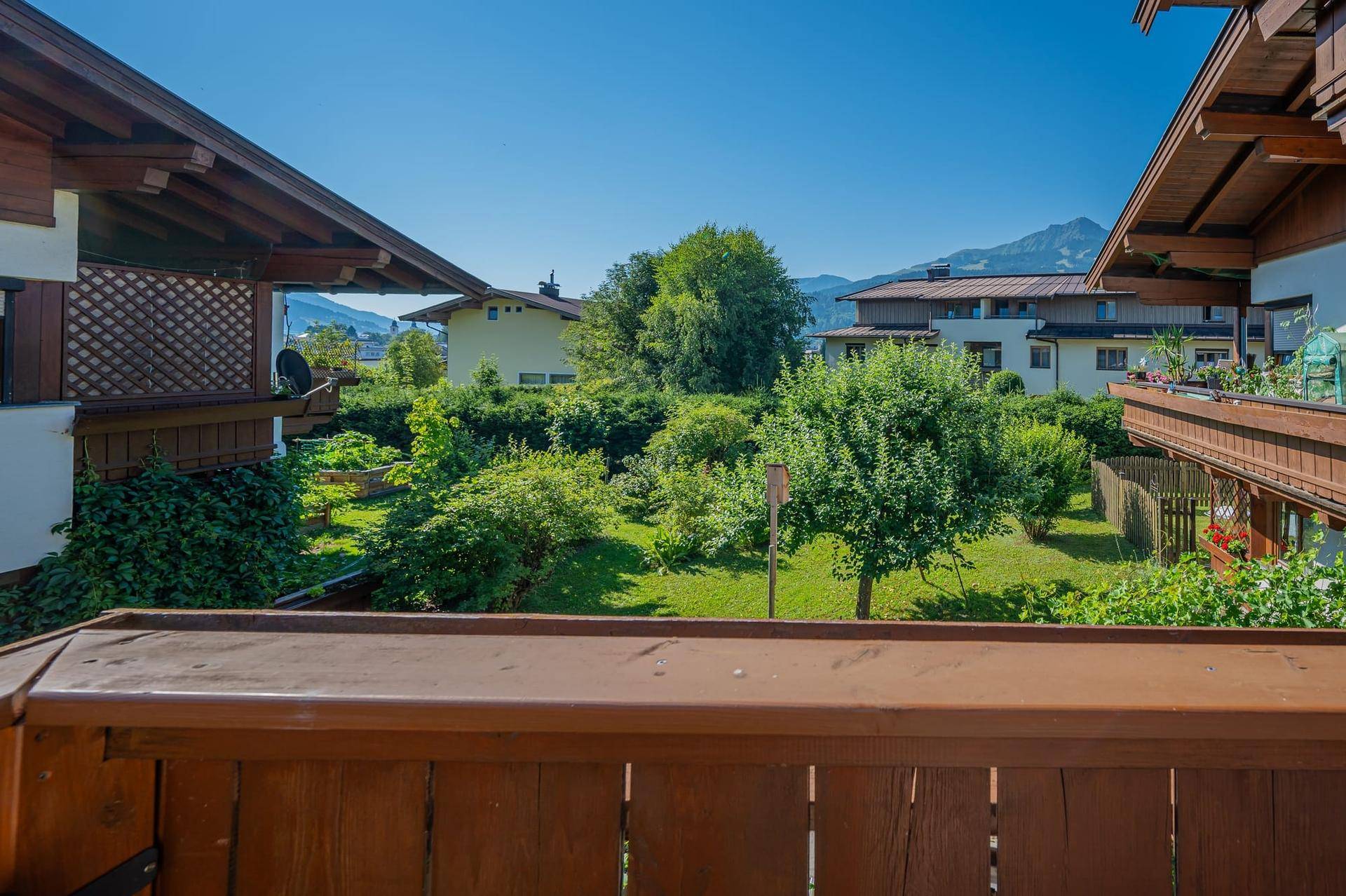 Blick von einem Balkon auf einen grünen Gartenbereich und die umliegende Berglandschaft.