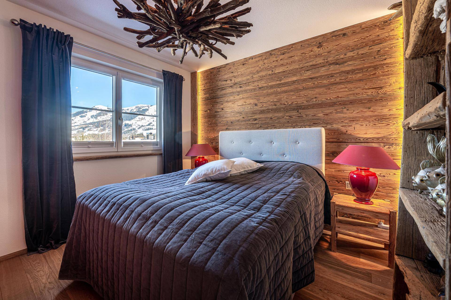 Schlafzimmer im alpinen Stil mit Holzwand, Doppelbett und Bergblick durch das Fenster.
