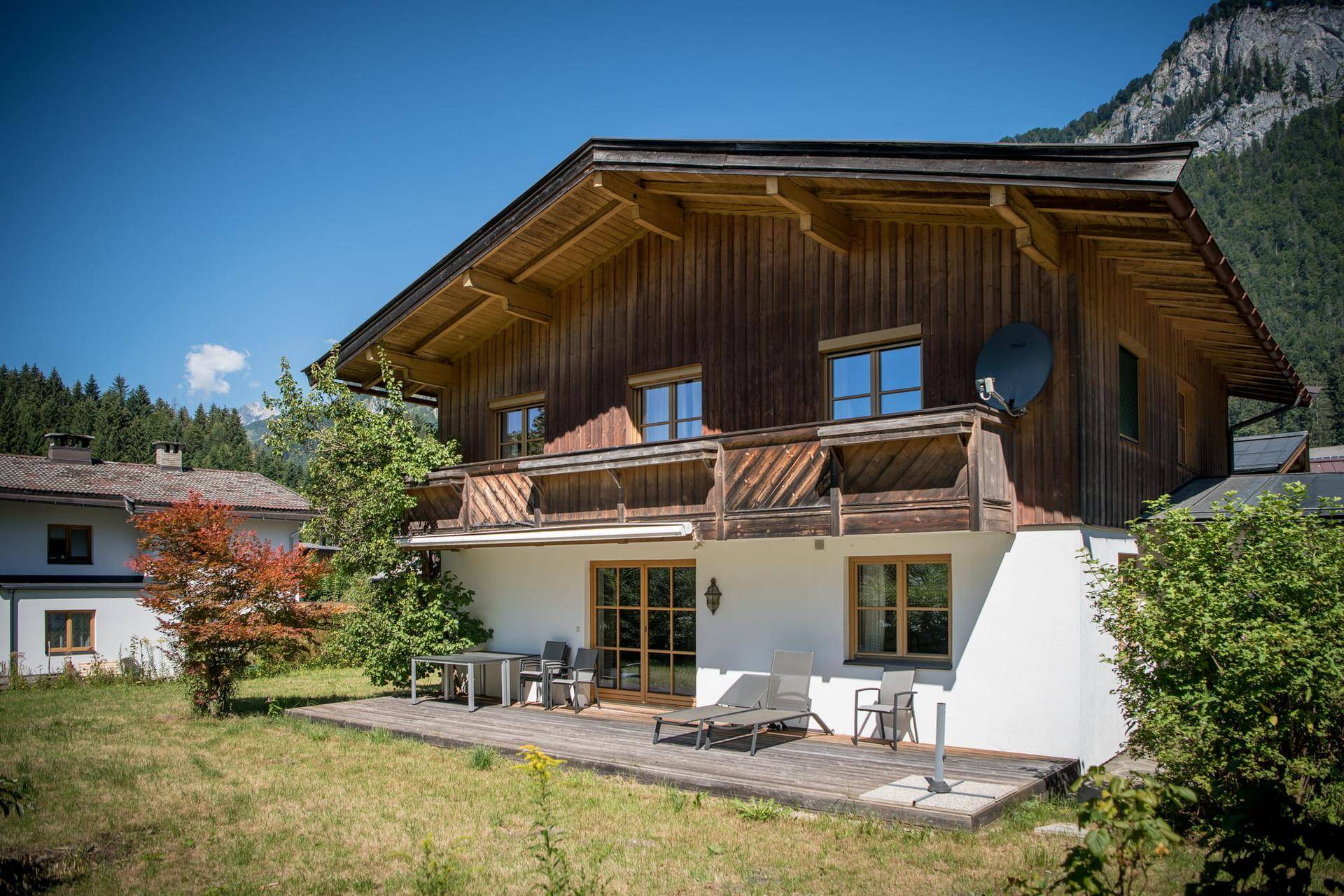 Holzhaus mit Terrasse und Bergblick, umgeben von grüner Natur unter blauem Himmel.