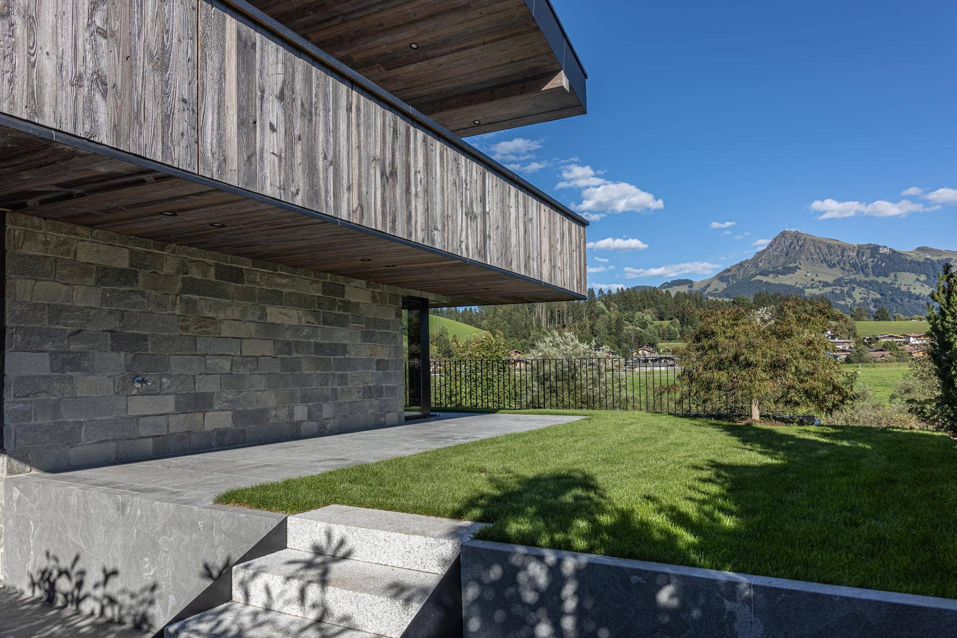 Moderne Architektur mit Steinfassade, gepflegtem Rasen und weitem Blick auf die umliegende Berglandschaft.