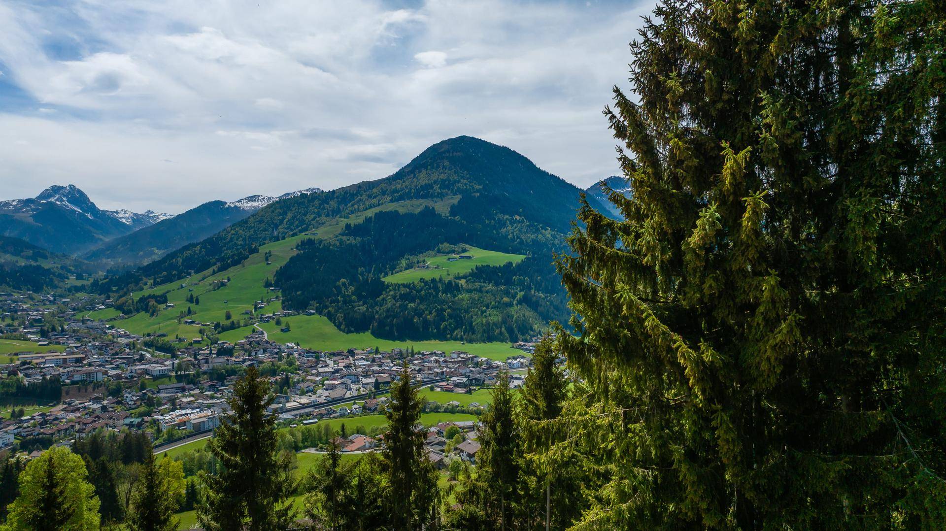 Panoramablick auf ein malerisches Tal mit einer Stadt, umgeben von grünen Hügeln und majestätischen Bergen.