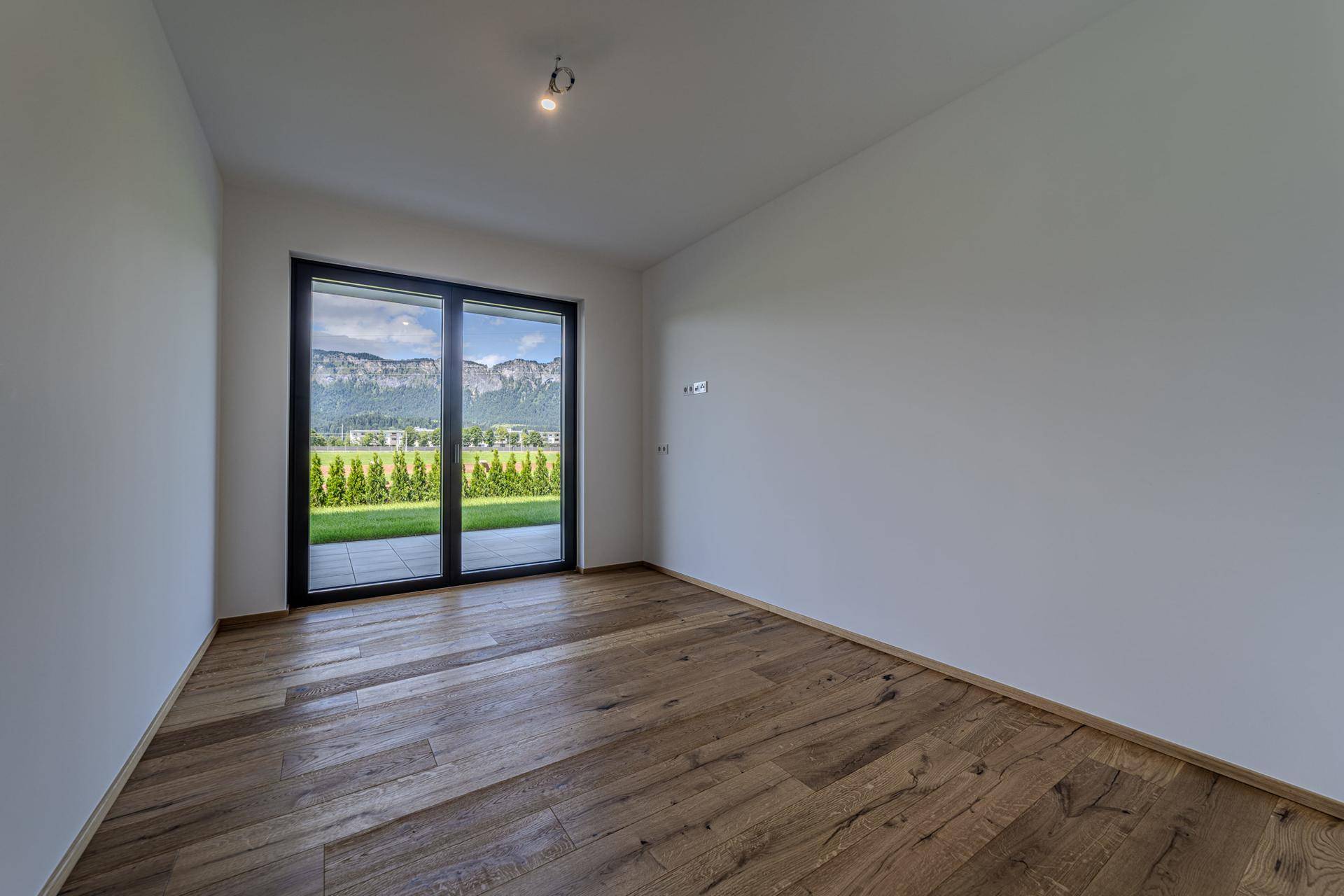 Leerer Raum mit Holzboden und großen Fenstern, die einen weiten Blick auf die Berge und den Garten bieten.